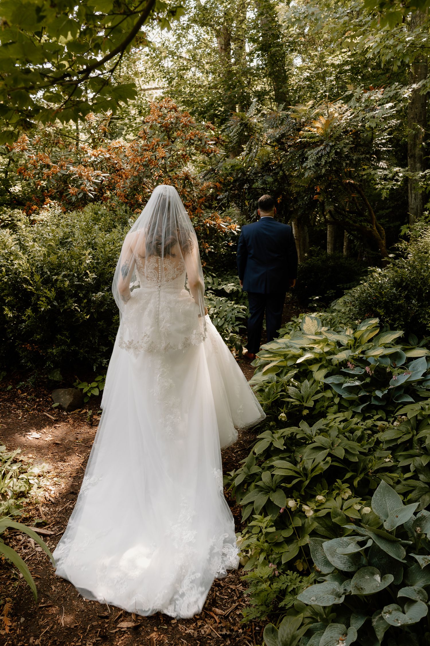 Bride in lace gown and veil walks with groom along a lush garden pathway.