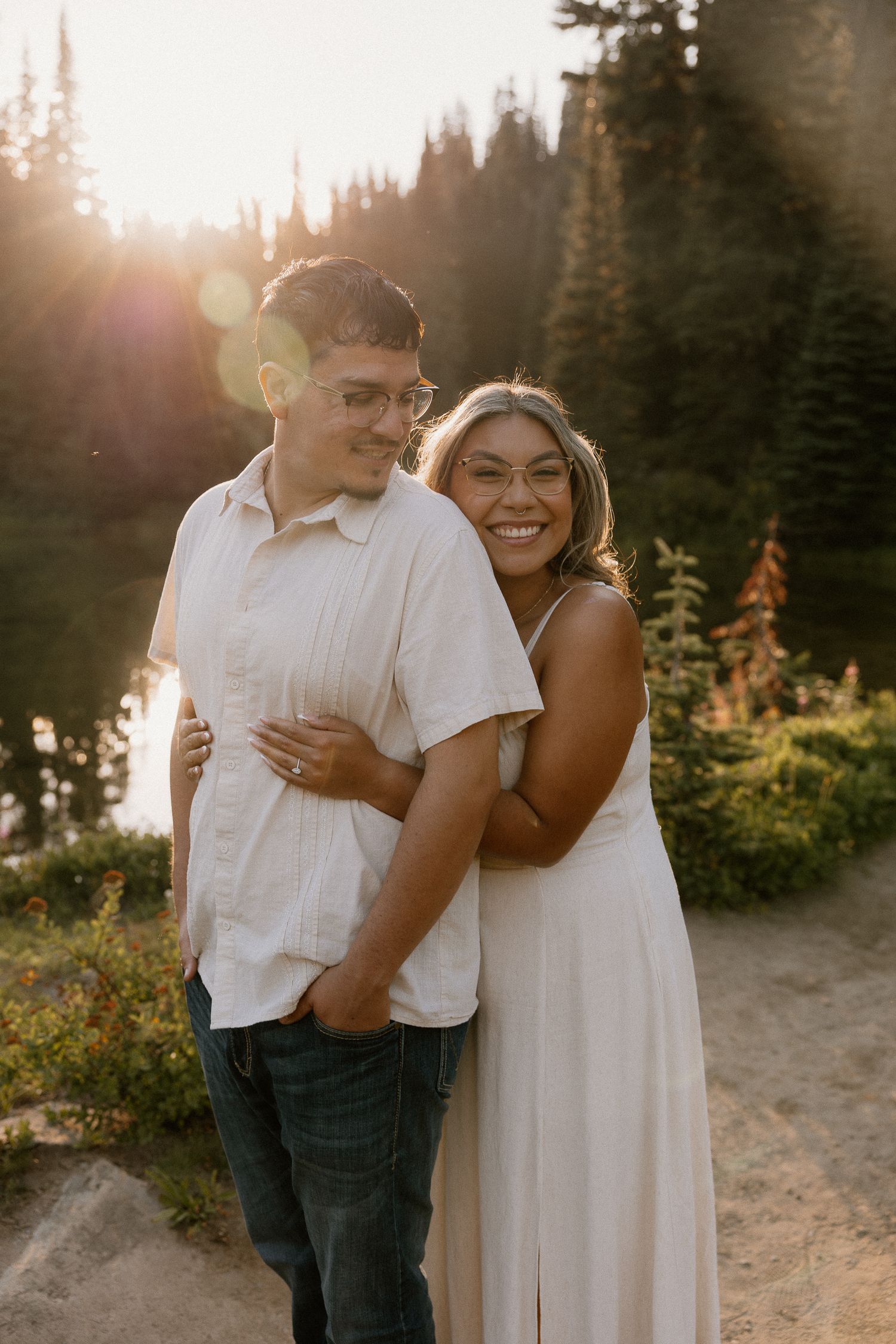 A couple embracing at golden hour near a mountain lake surrounded by evergreen trees.