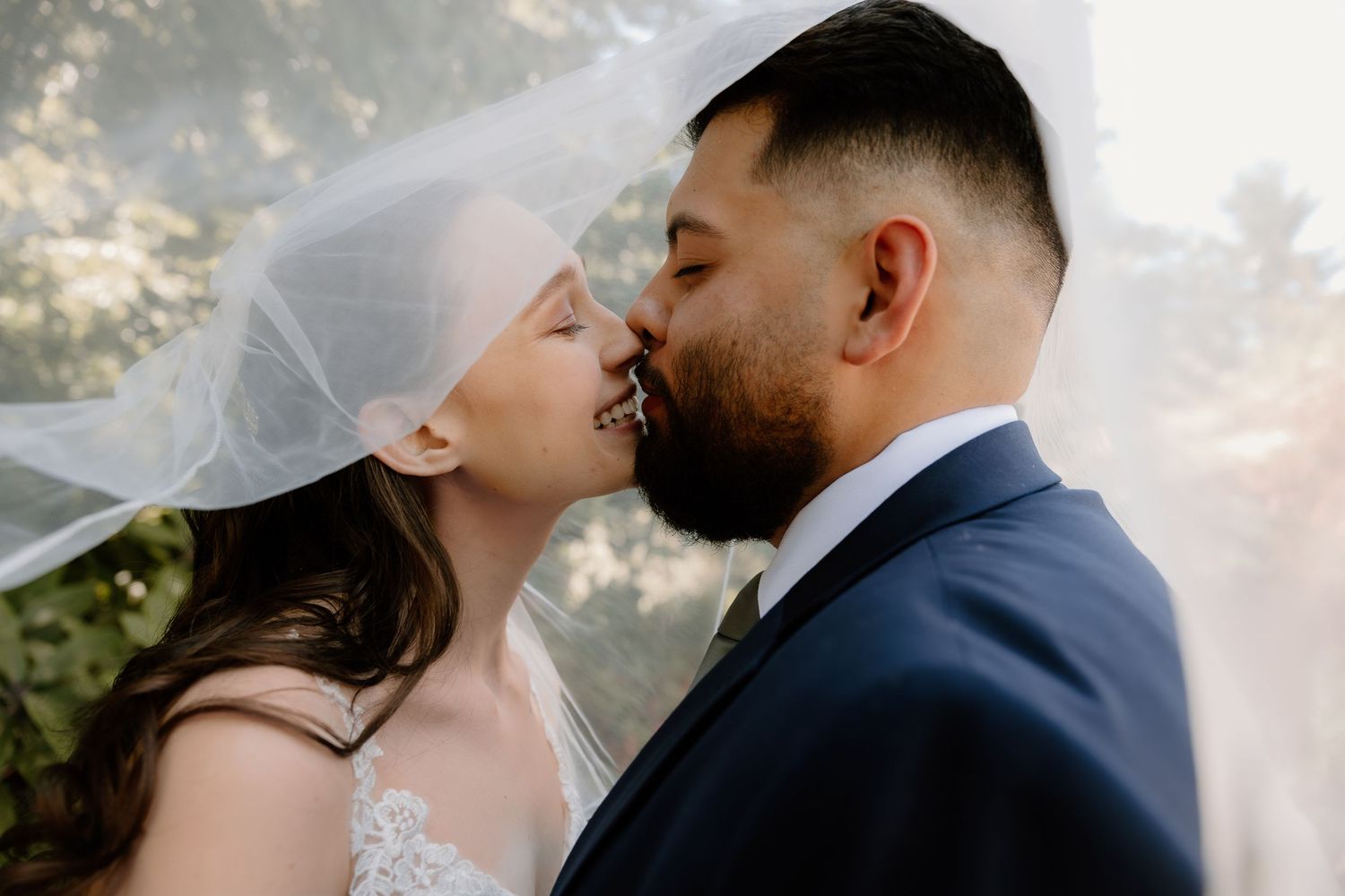 Bride and groom sharing a romantic kiss under a flowing white wedding veil outdoors.