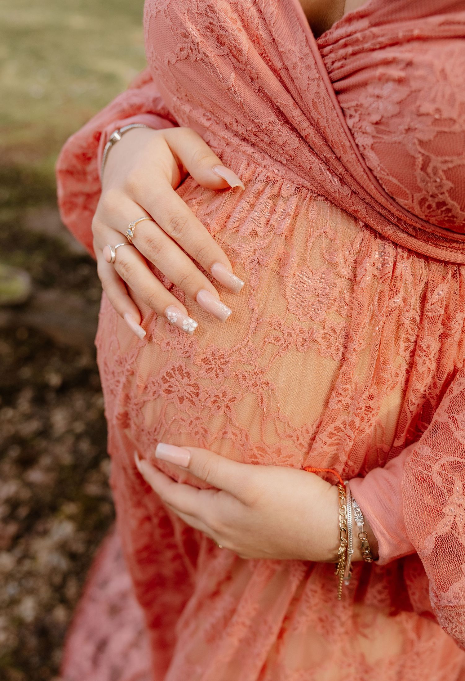 A pregnant woman in a coral lace dress cradles her baby bump with manicured hands and gold bracelets.