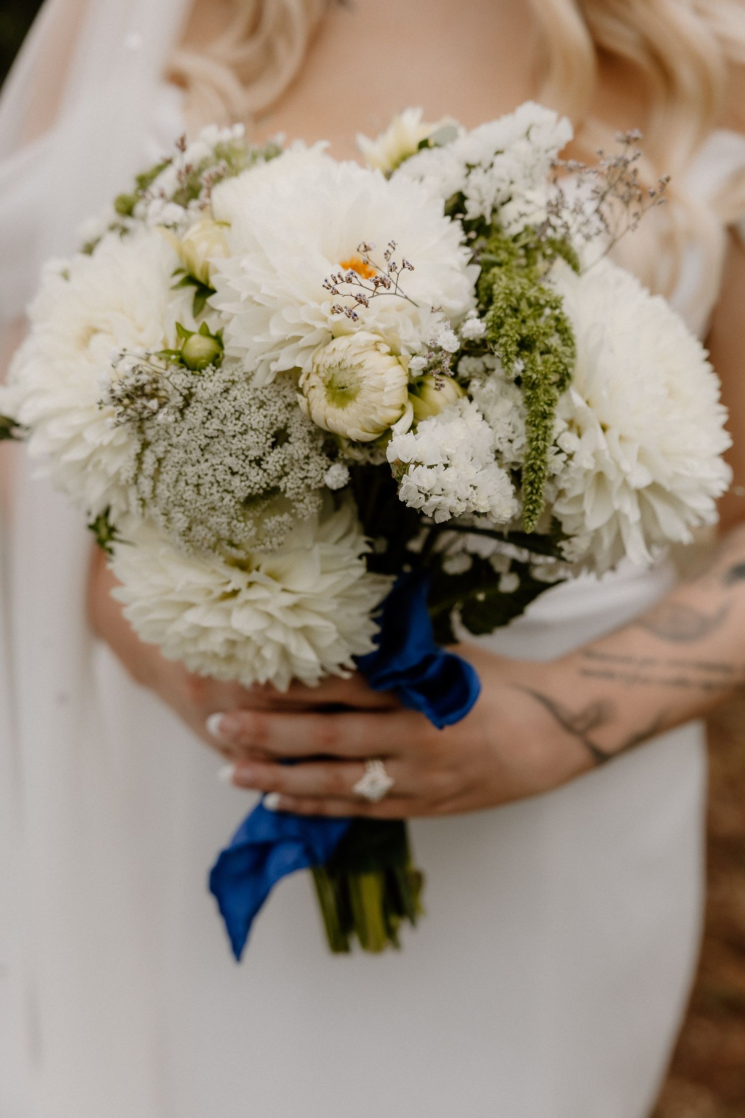 Bride holds a stunning white wedding bouquet with dahlias and greenery, tied with a royal blue ribbon.