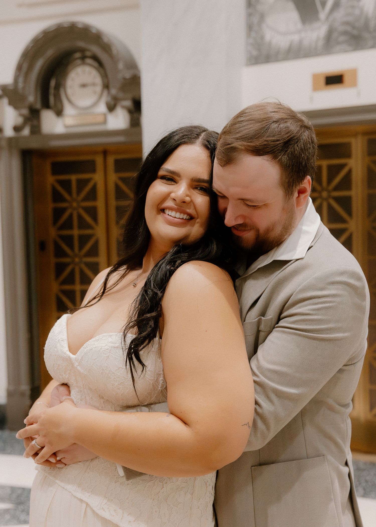 Groom embraces bride from behind as she smiles brightly outside ornate courthouse doors.