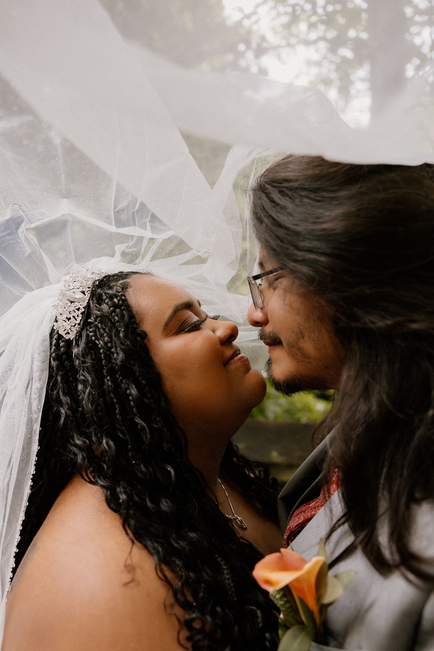Bride and groom share intimate moment under flowing white veil surrounded by soft natural light.