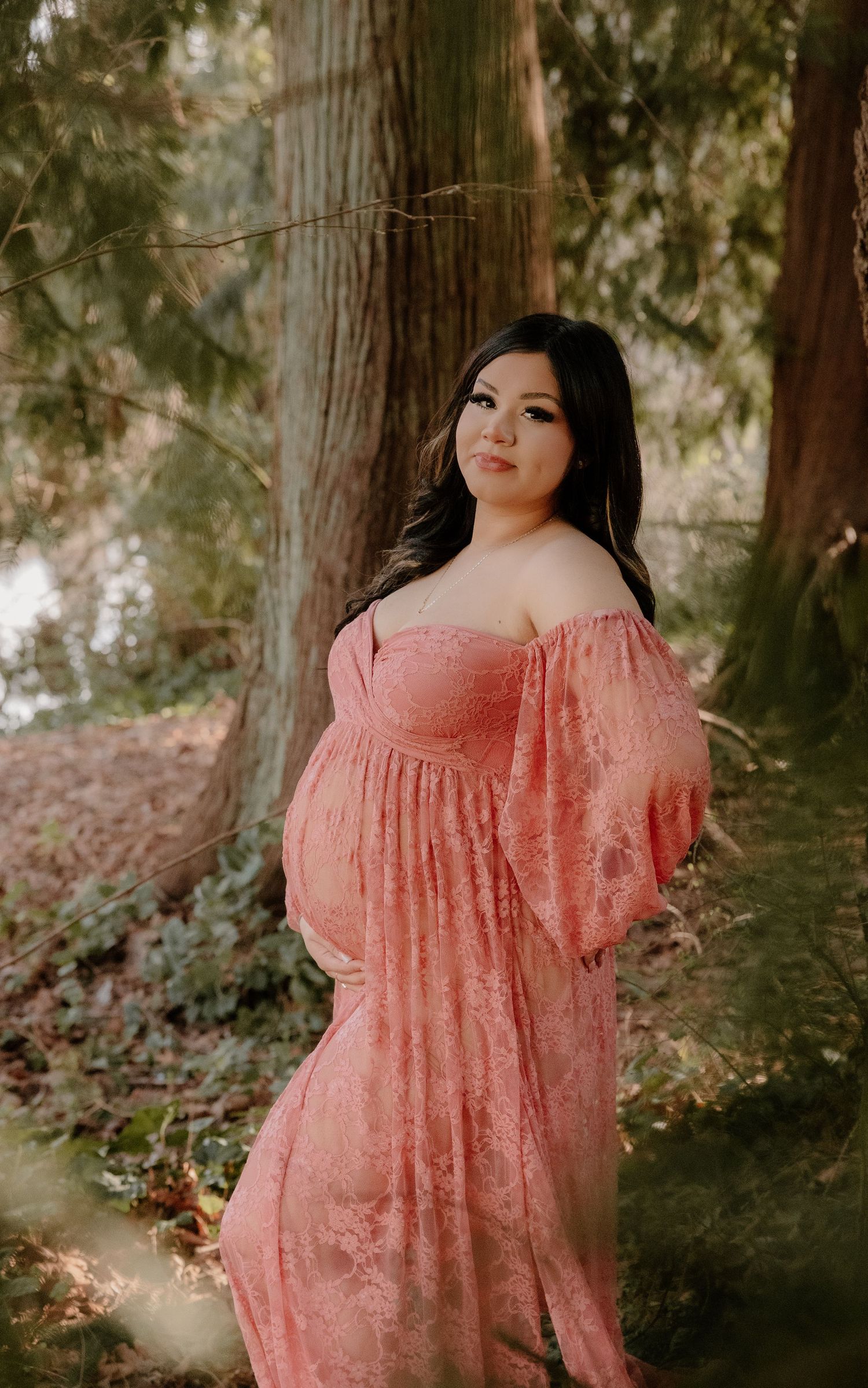 Pregnant woman in pink lace off-shoulder maternity gown posing outdoors among tall trees in a forest setting.