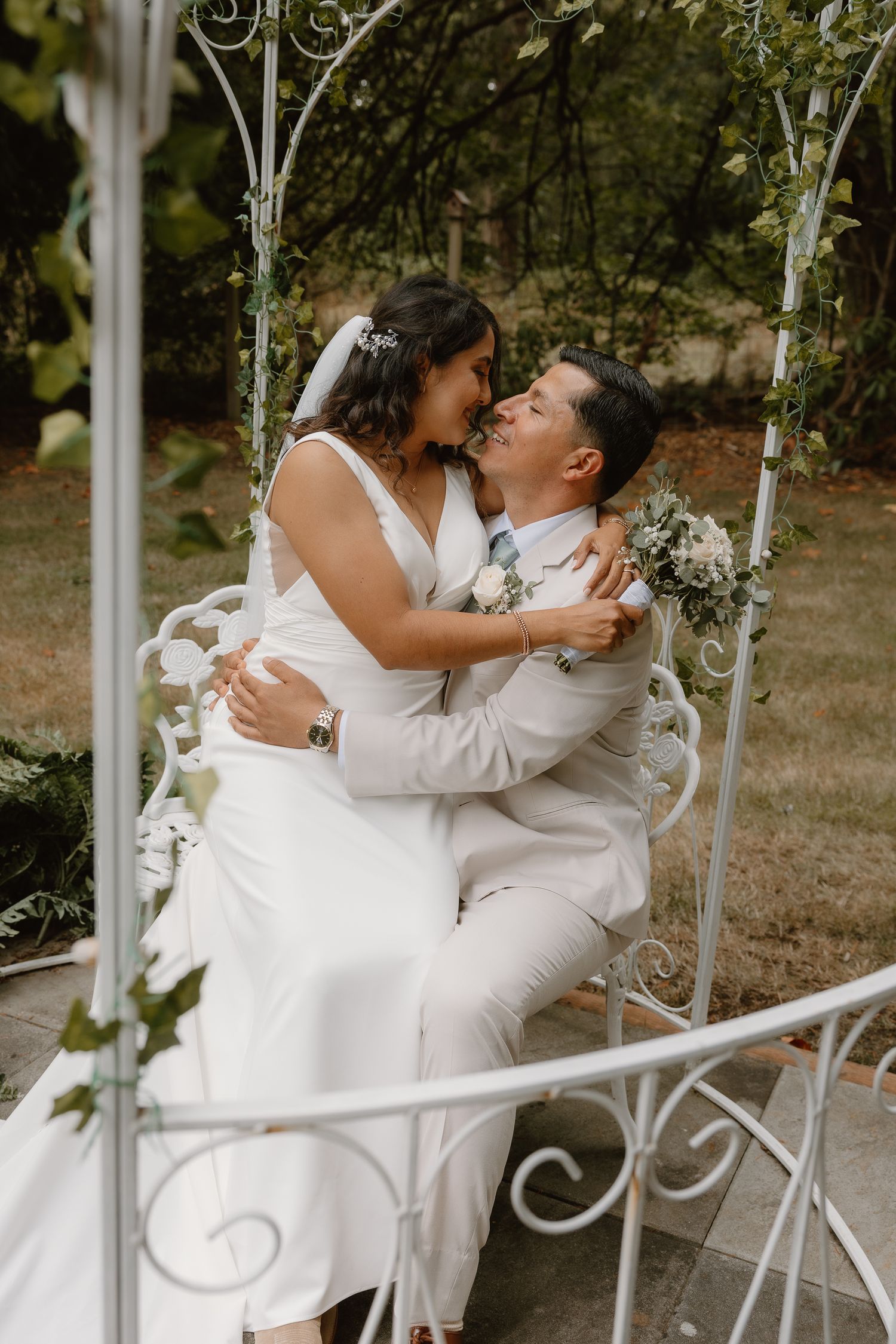 Bride and groom sharing a kiss while seated on white iron bench under garden arbor.