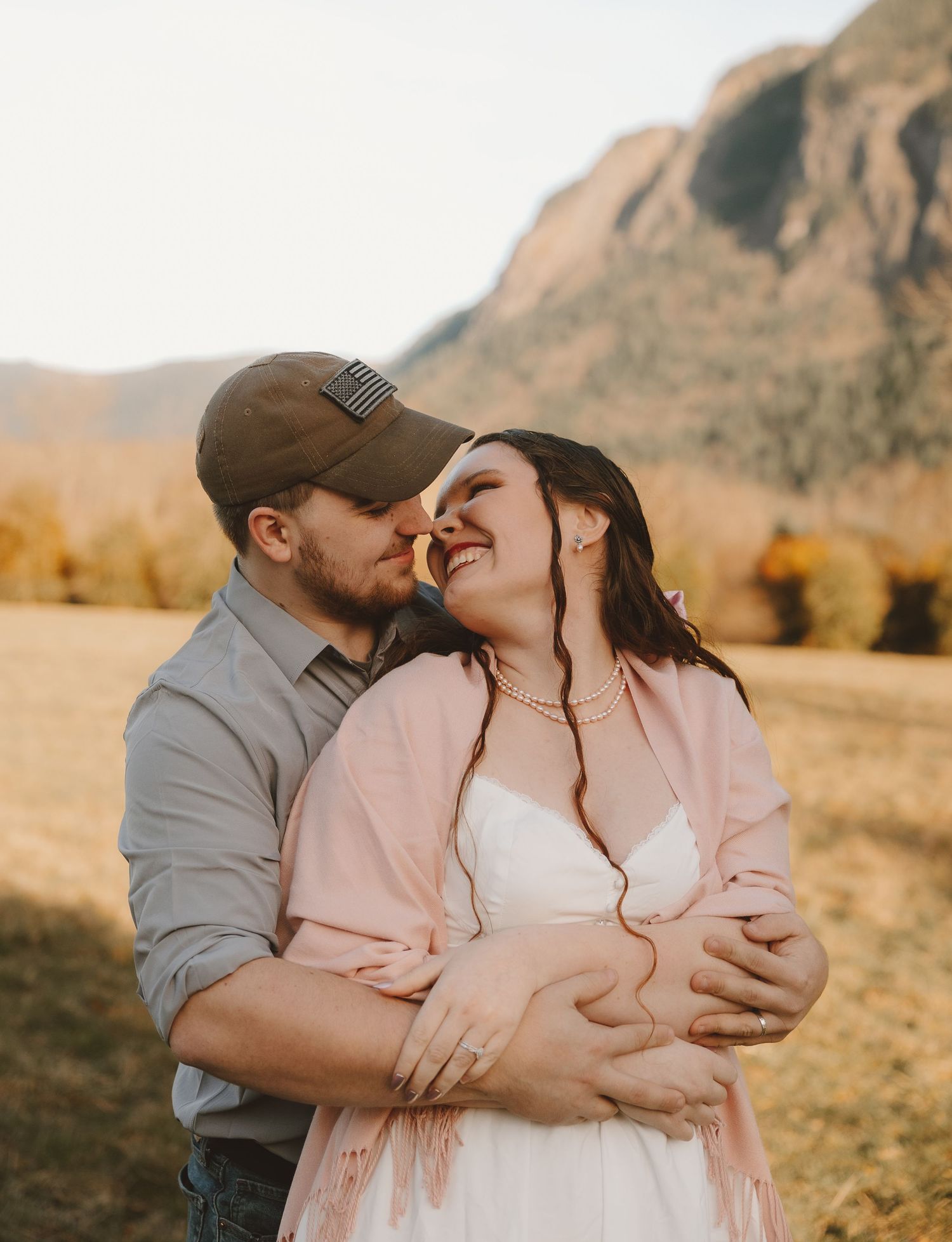 A couple shares a romantic moment in a golden mountain meadow, the man kissing her cheek as she smiles joyfully.