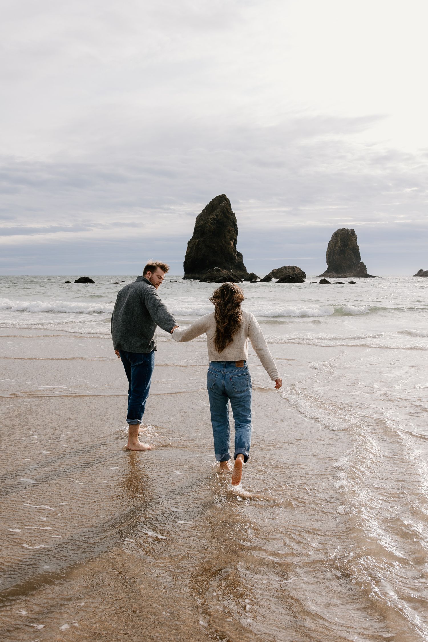 Couple holding hands walking barefoot in ocean waves at Cannon Beach with iconic sea stacks in background.