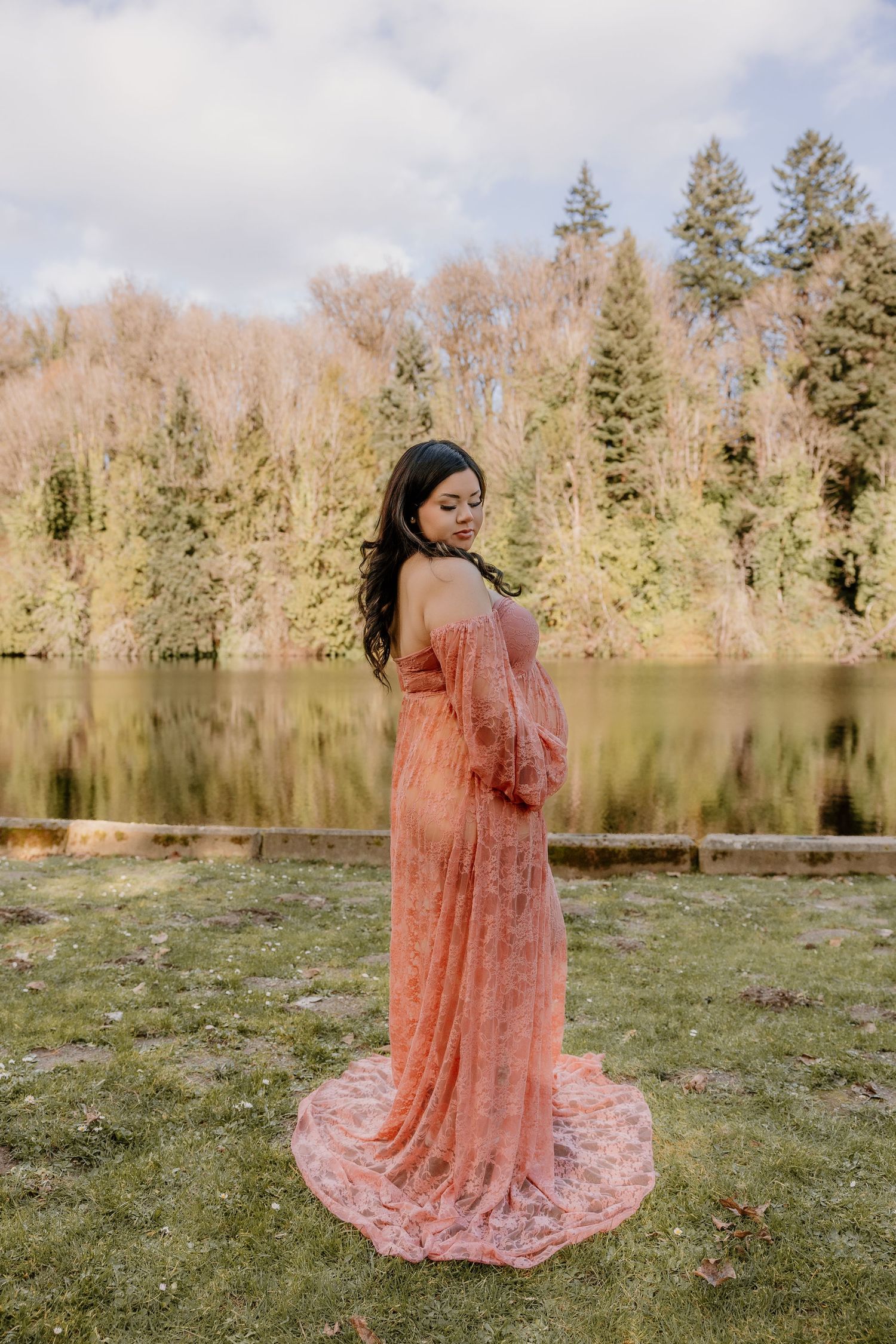 Pregnant woman in flowing pink lace gown standing elegantly by a calm lake surrounded by trees.