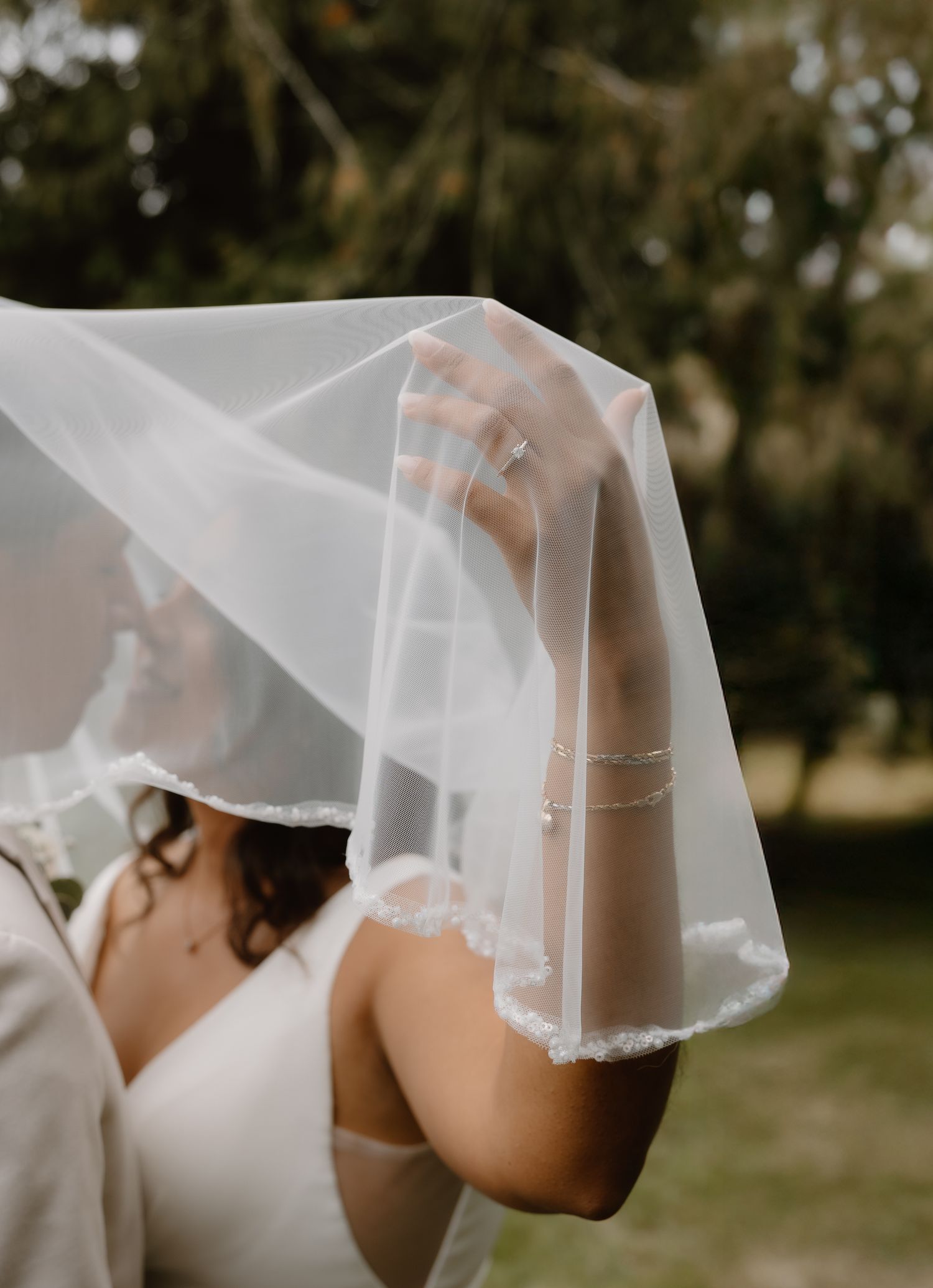Close-up of bride lifting delicate white wedding veil outdoors showing diamond engagement ring.