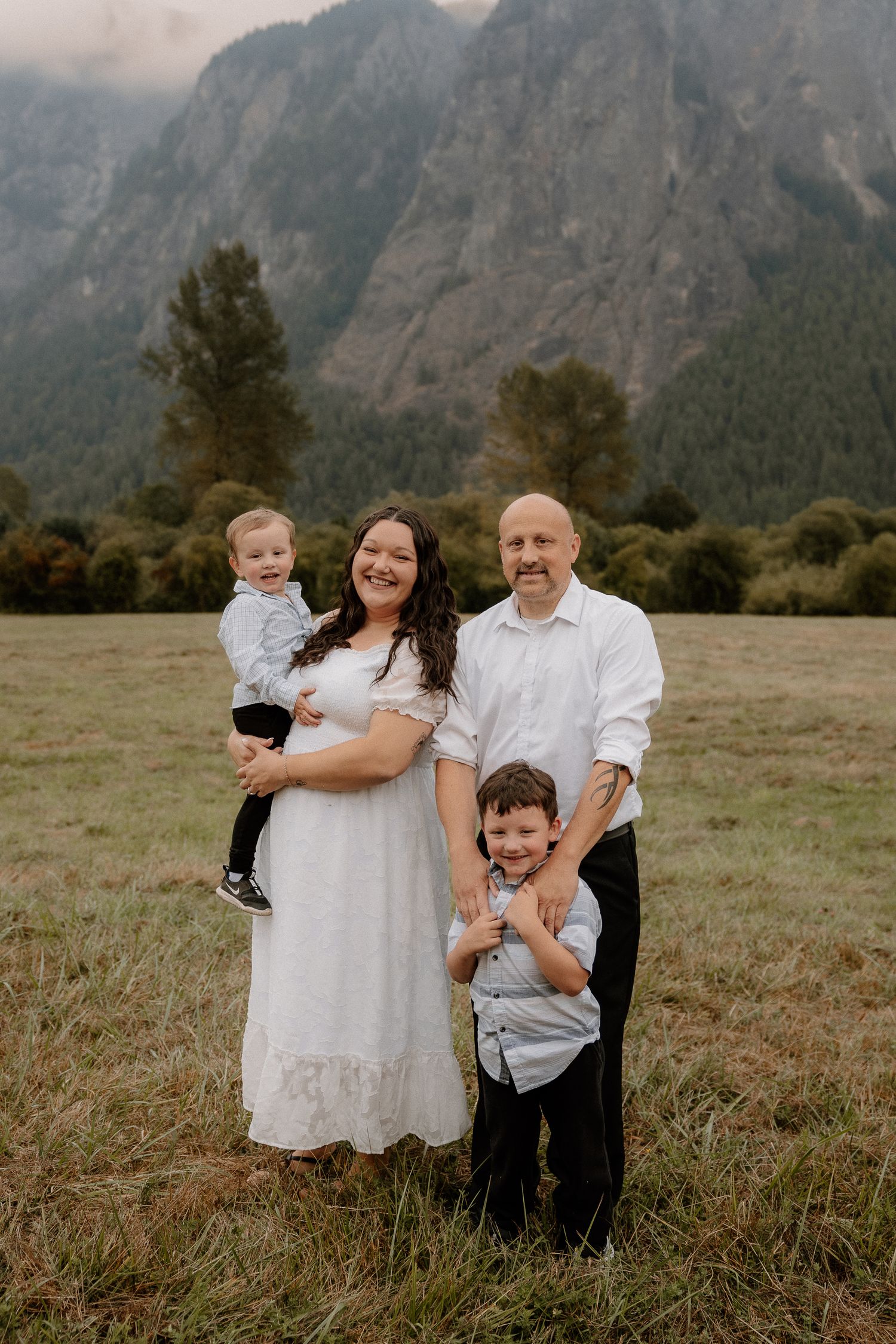 Smiling family of four poses in grassy meadow with majestic cloudy mountain range behind them.
