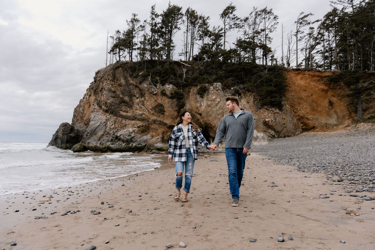 Couple holding hands walking on a rocky beach with dramatic cliffs and evergreen trees in the background.