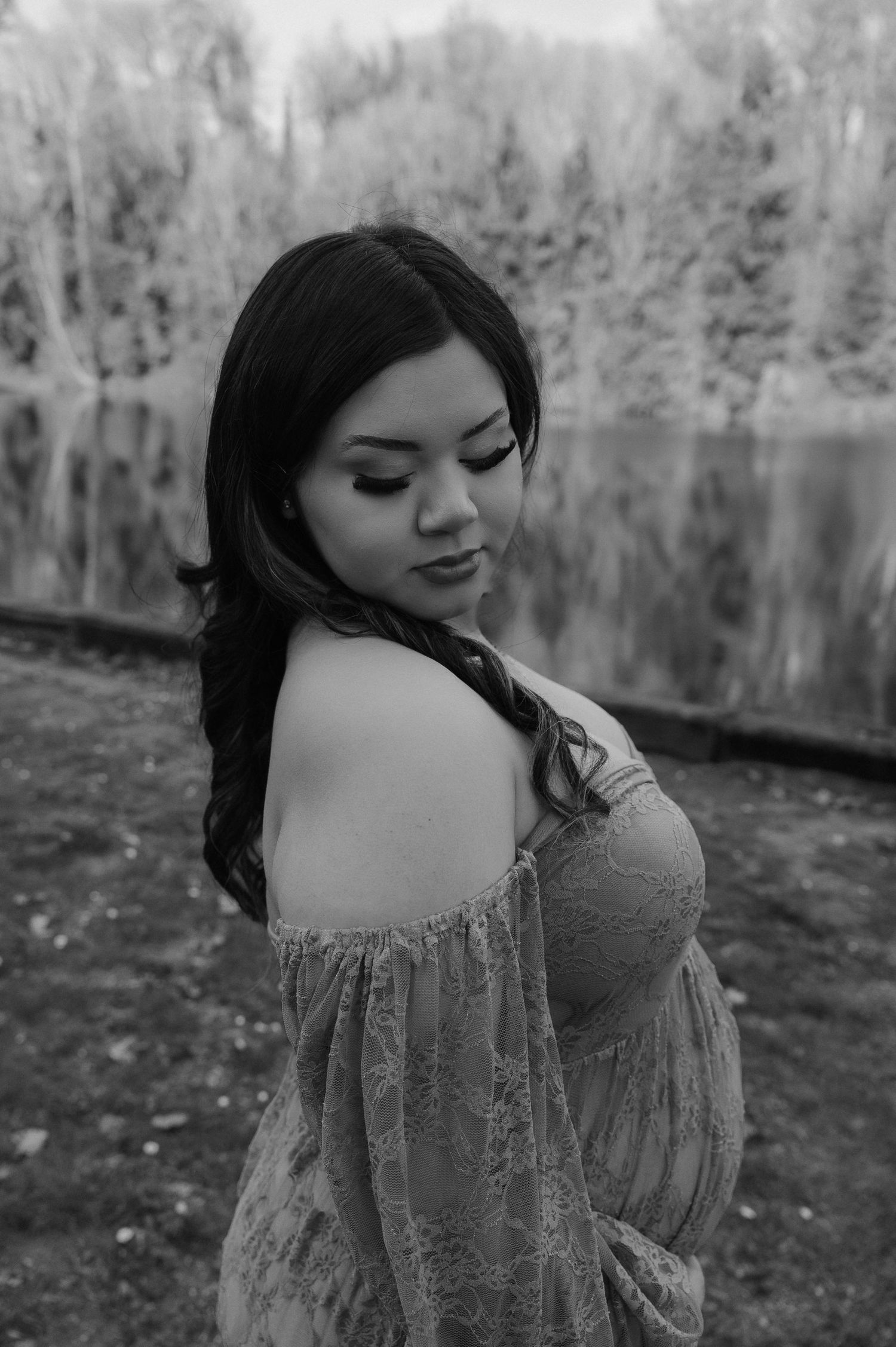Black and white maternity portrait of woman in lace dress showing bare shoulder near reflective lake.