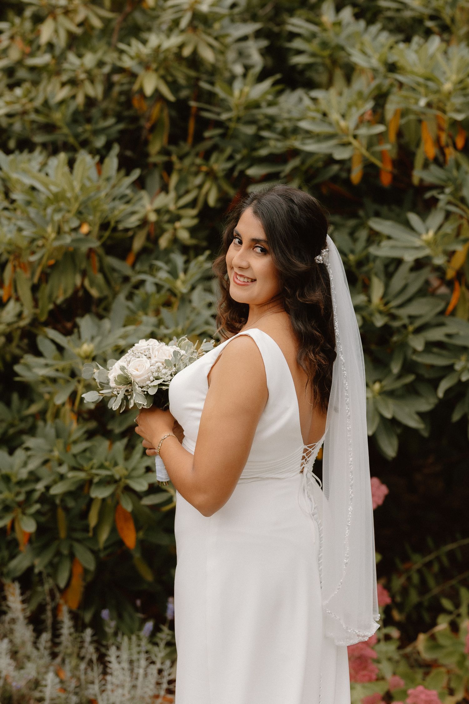 Smiling bride in white sleeveless gown holding white bouquet with veil before green foliage.