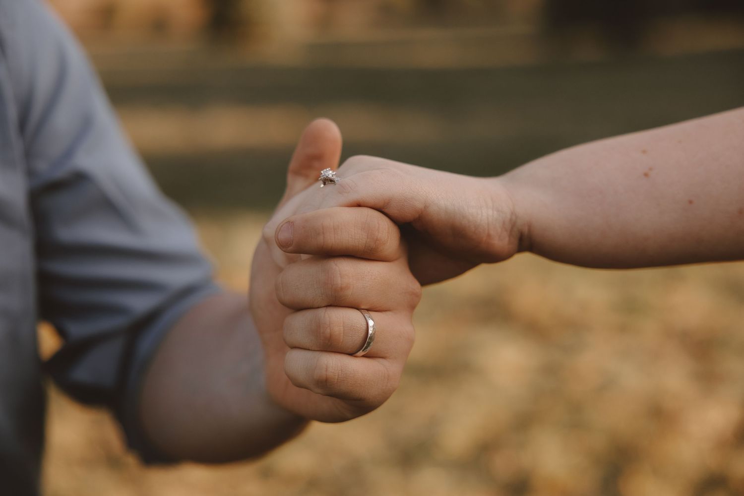 Close-up of two hands touching, one wearing a silver wedding ring, with a blurred outdoor background.