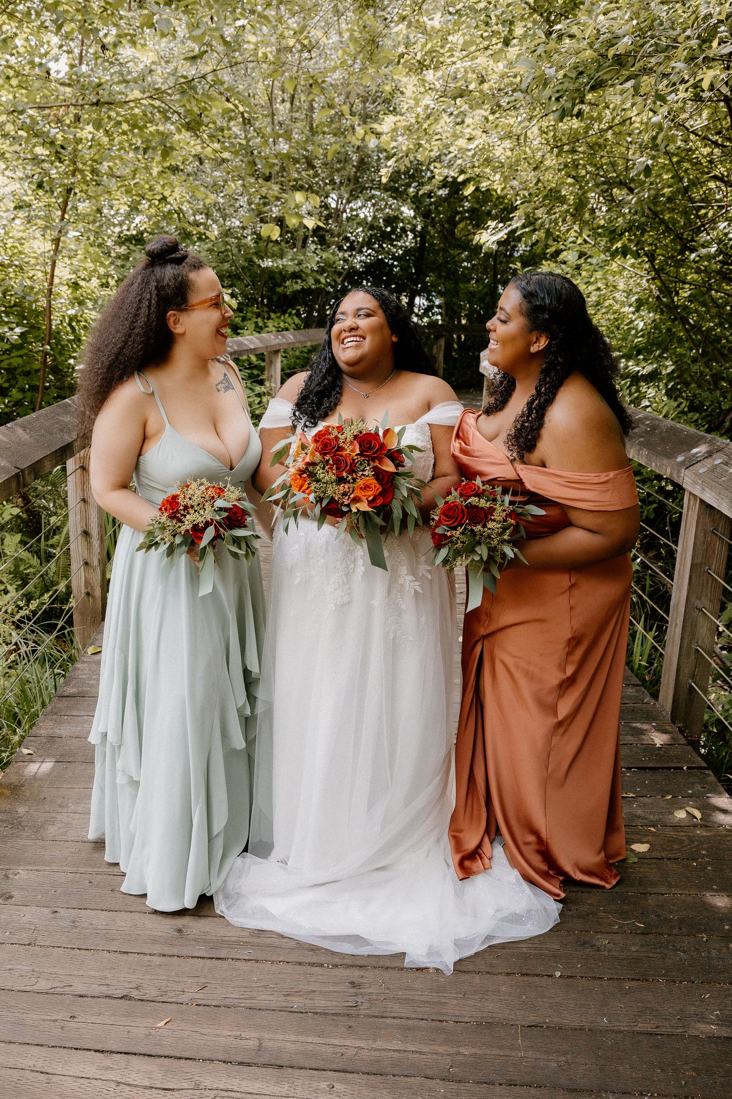 A bride in white stands with two bridesmaids in sage and rust gowns holding vibrant fall bouquets.