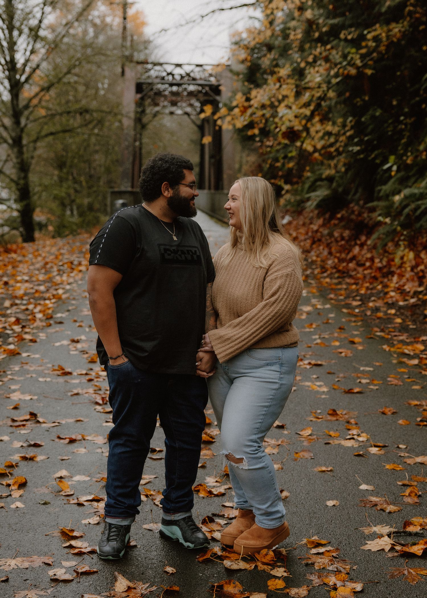 A couple holding hands and laughing together on a leaf-covered path with an iron bridge behind them.