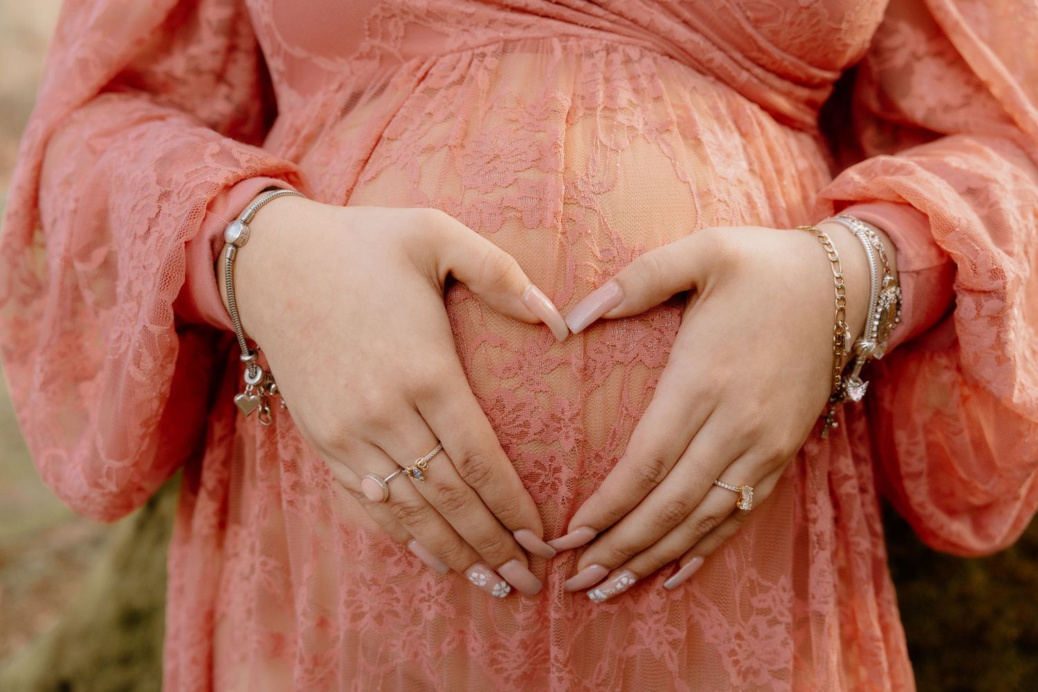 Close-up of pregnant woman making heart shape with hands over baby bump in pink lace dress.