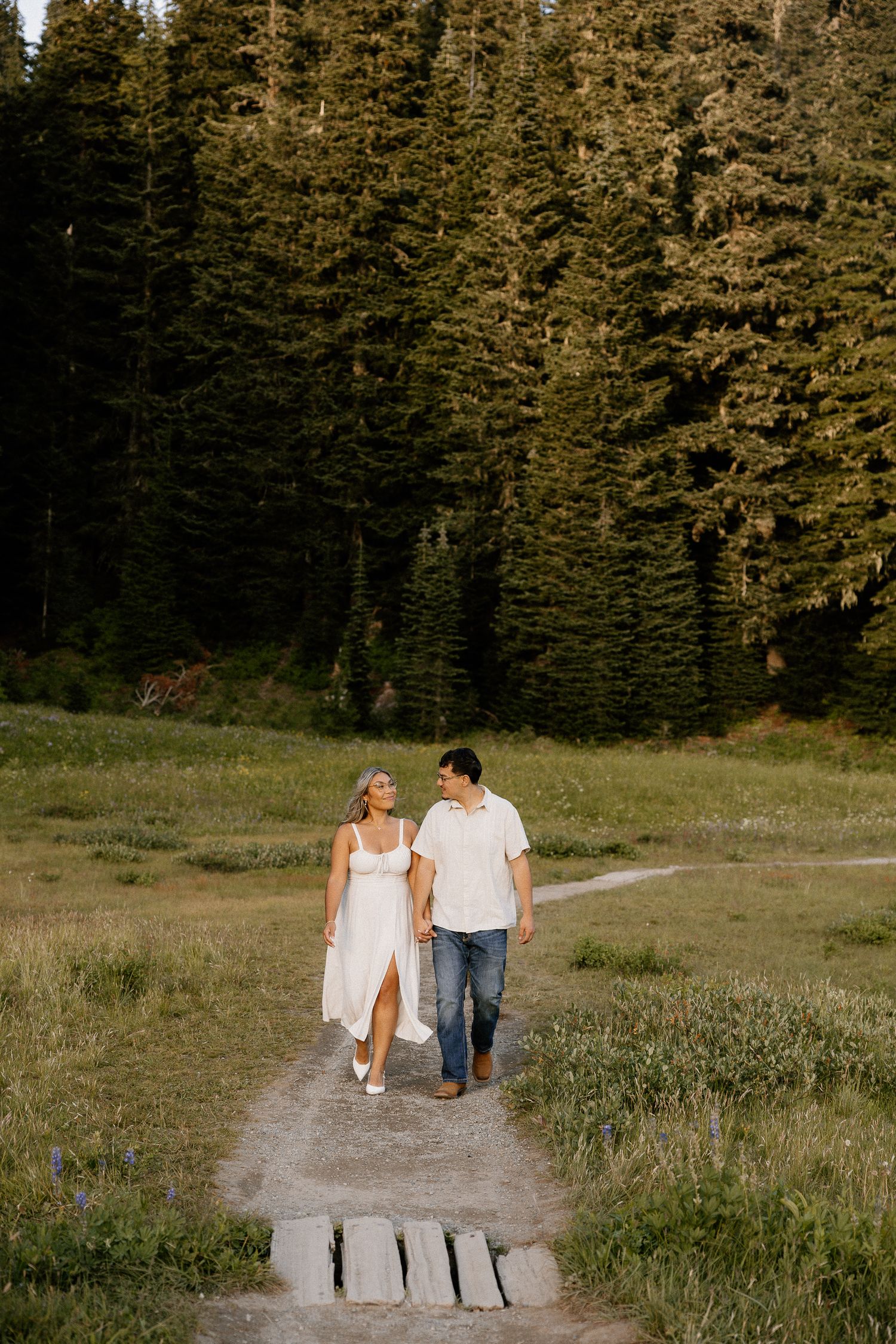 A couple walking hand in hand along a trail through a lush green mountain meadow.