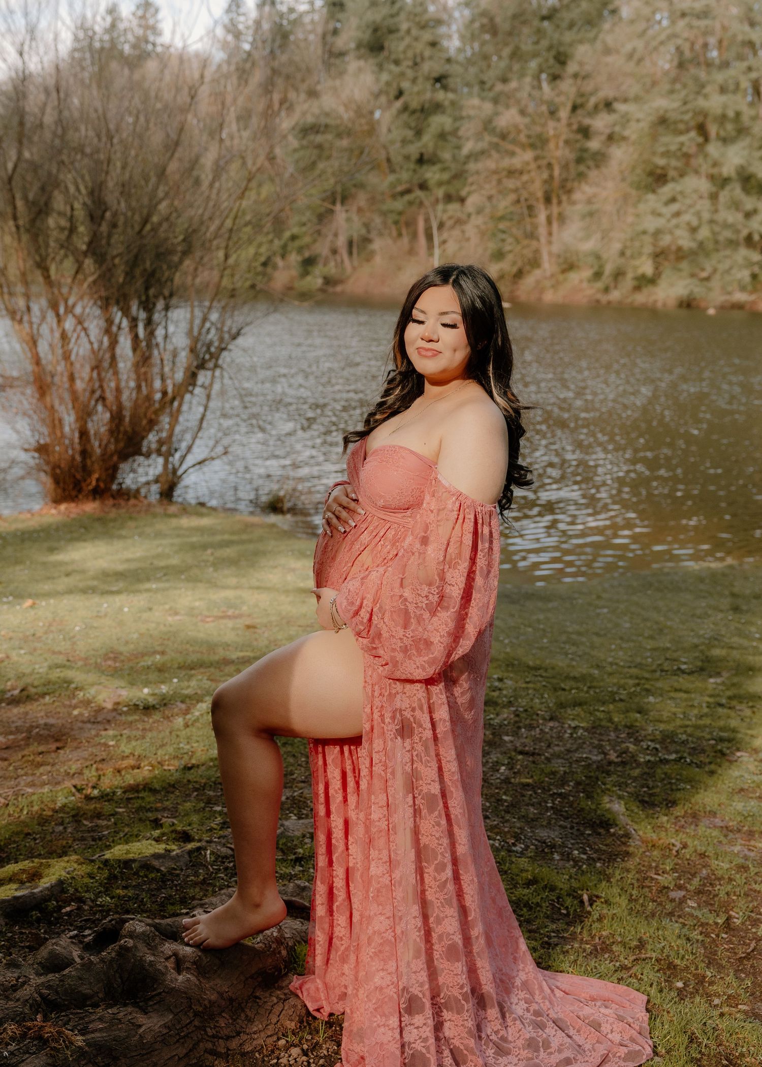 Expecting mother in a pink lace maternity dress posing by a serene lake with her baby bump.