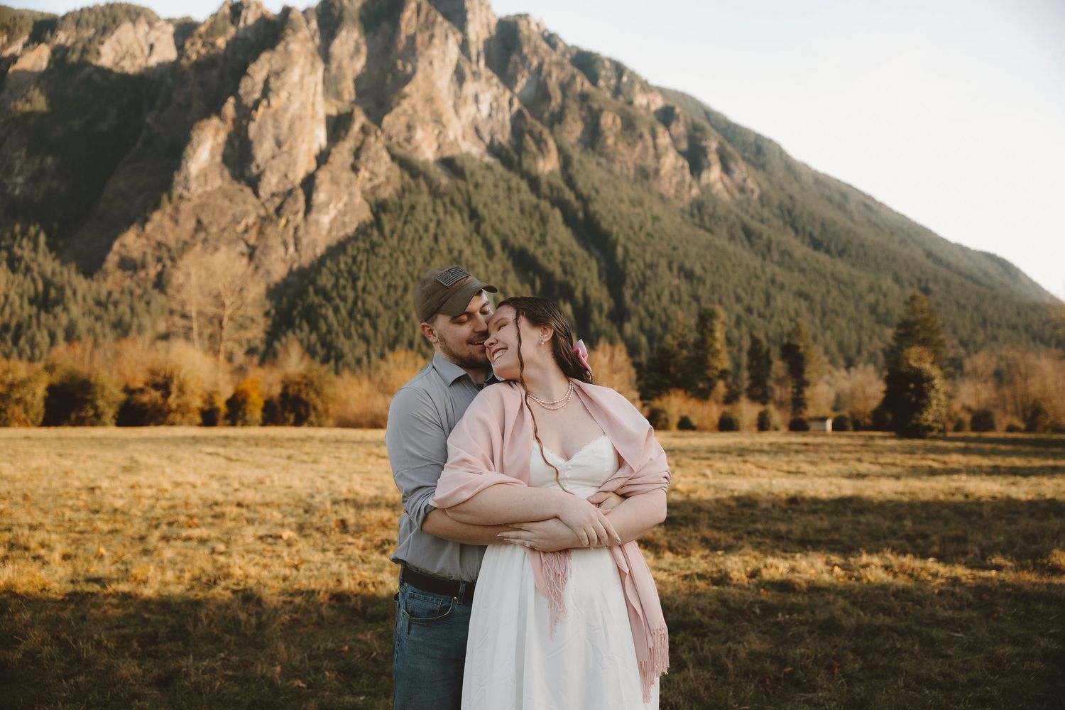 Couple embracing in golden field with mountain backdrop during outdoor engagement photo session.