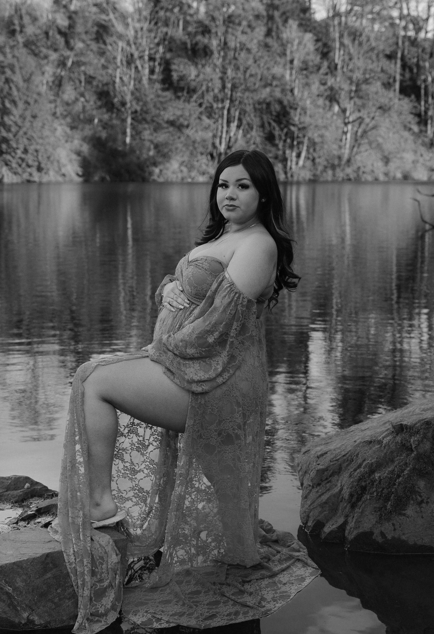Black and white maternity photo of a woman in lace gown posing on rocks by a serene lake surrounded by trees.