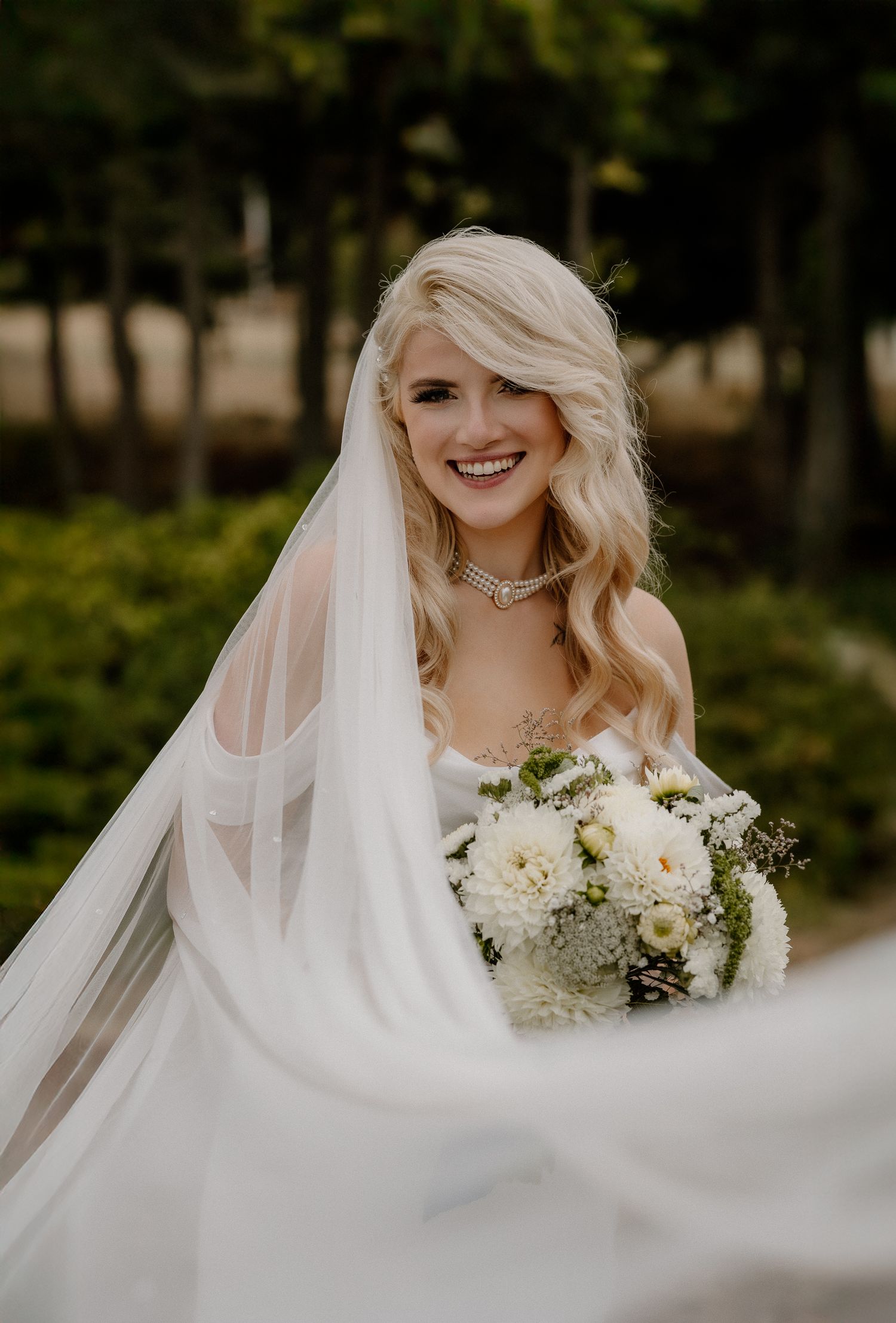 Radiant blonde bride smiles in a white gown and veil, holding a white floral bouquet in a garden.