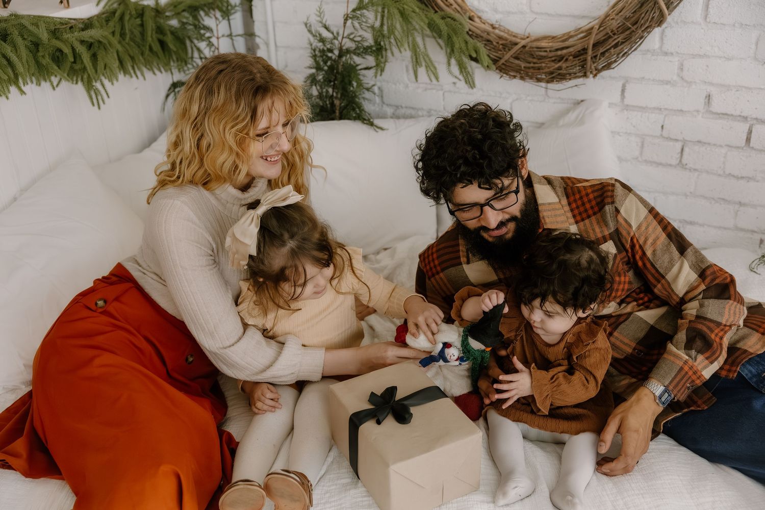 Family of four sitting together on a white couch opening Christmas gifts in a festive room.