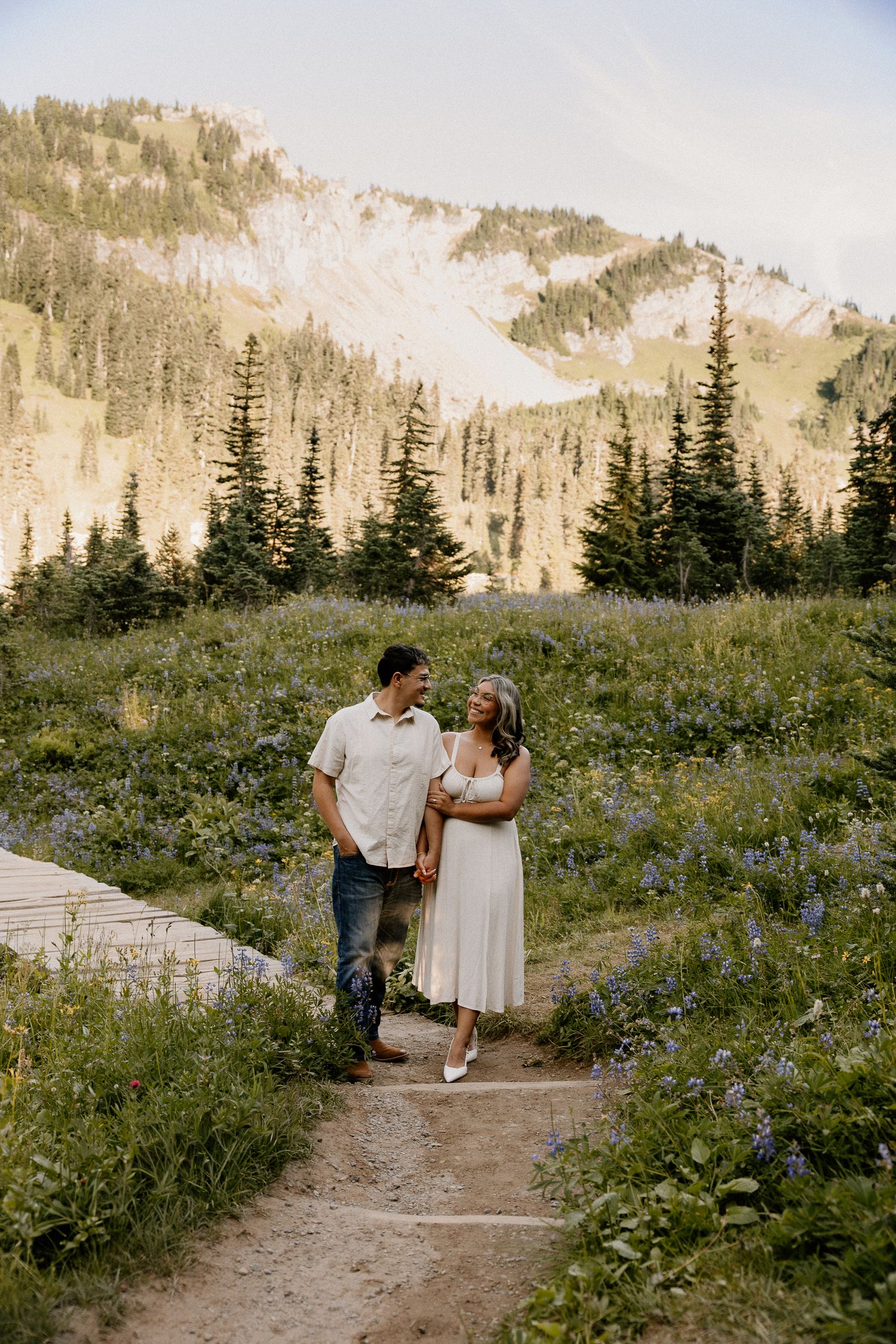 A couple laughing together on a path through a wildflower meadow with a mountain backdrop.