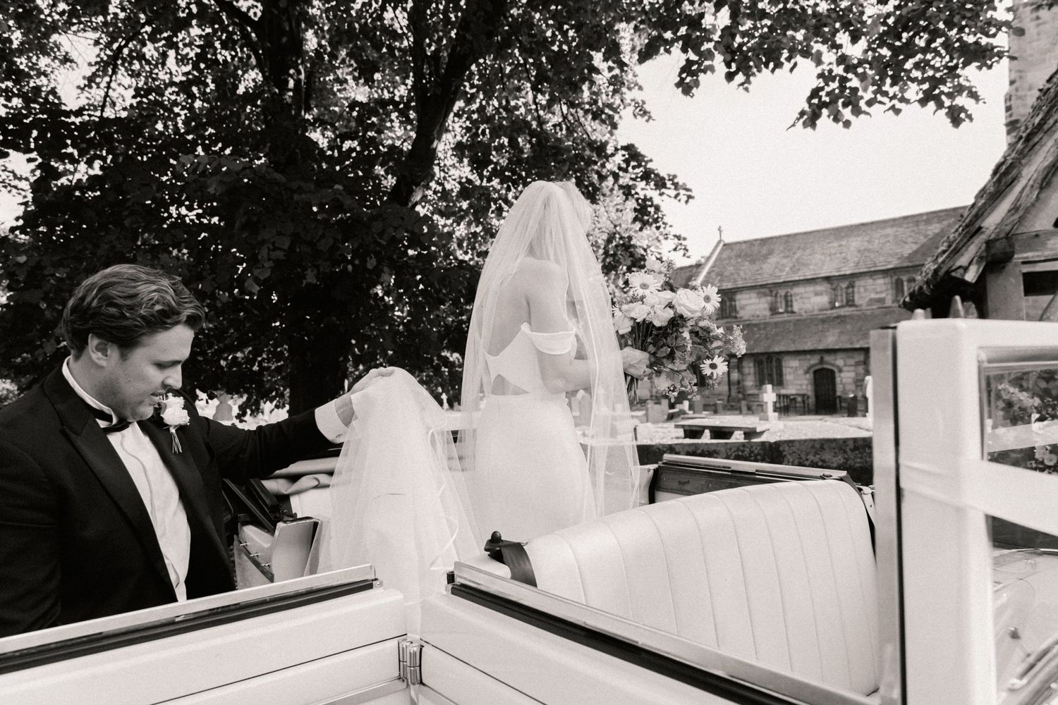 Groom holding brides wedding dress helping her get into vintage car outside St Wilfrids church