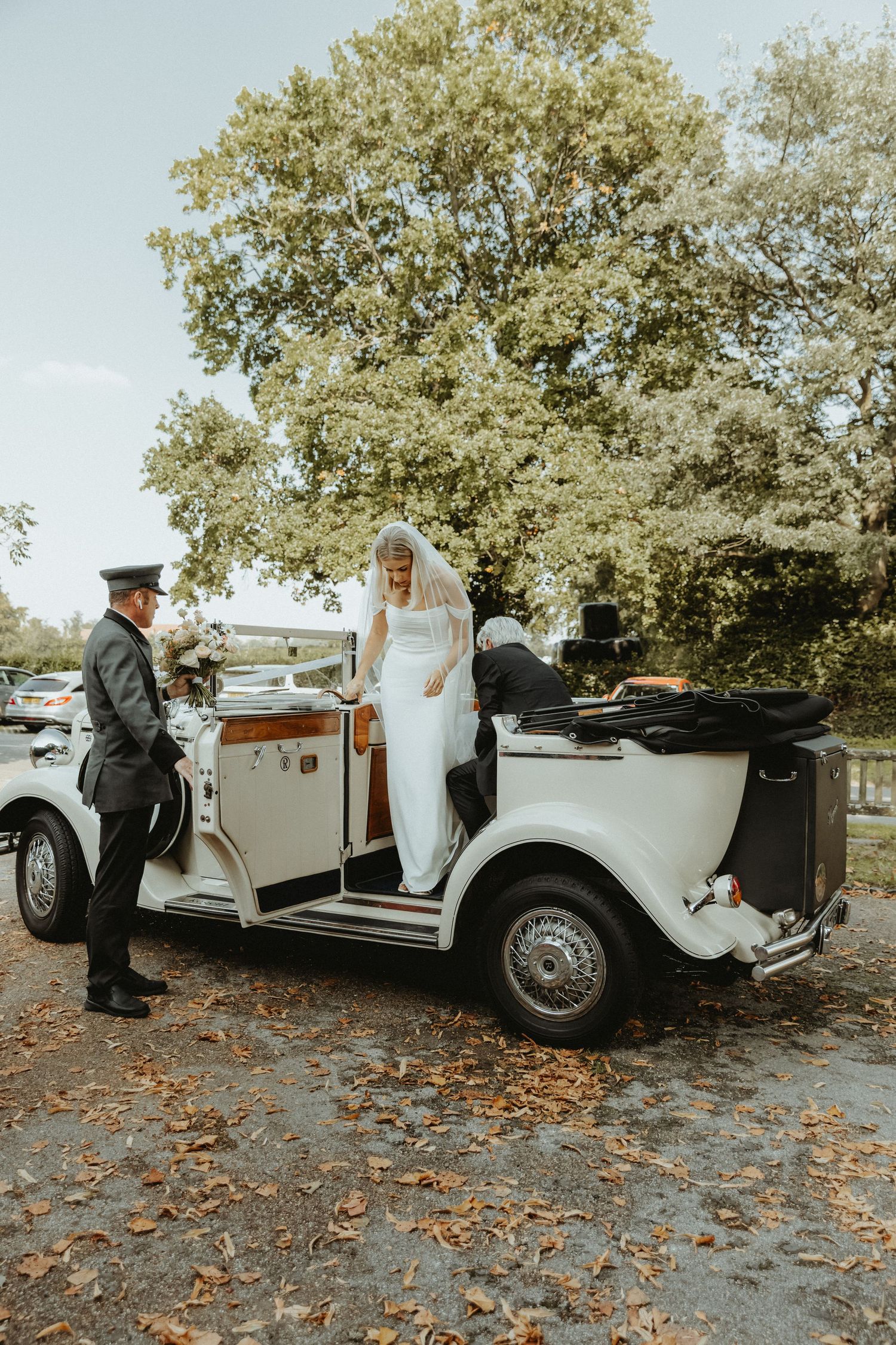 Bride getting out of vintage wedding car with roof down outside of St Wilfrids church Mobberley