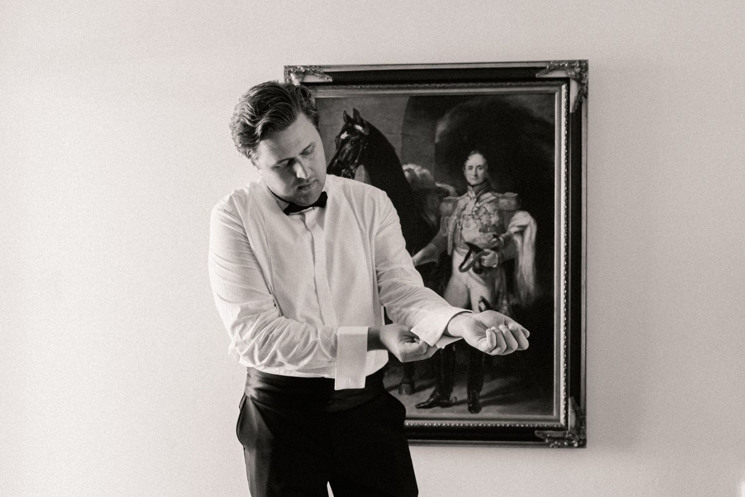 Black and white image of groom putting cufflinks on with oil painting behind him