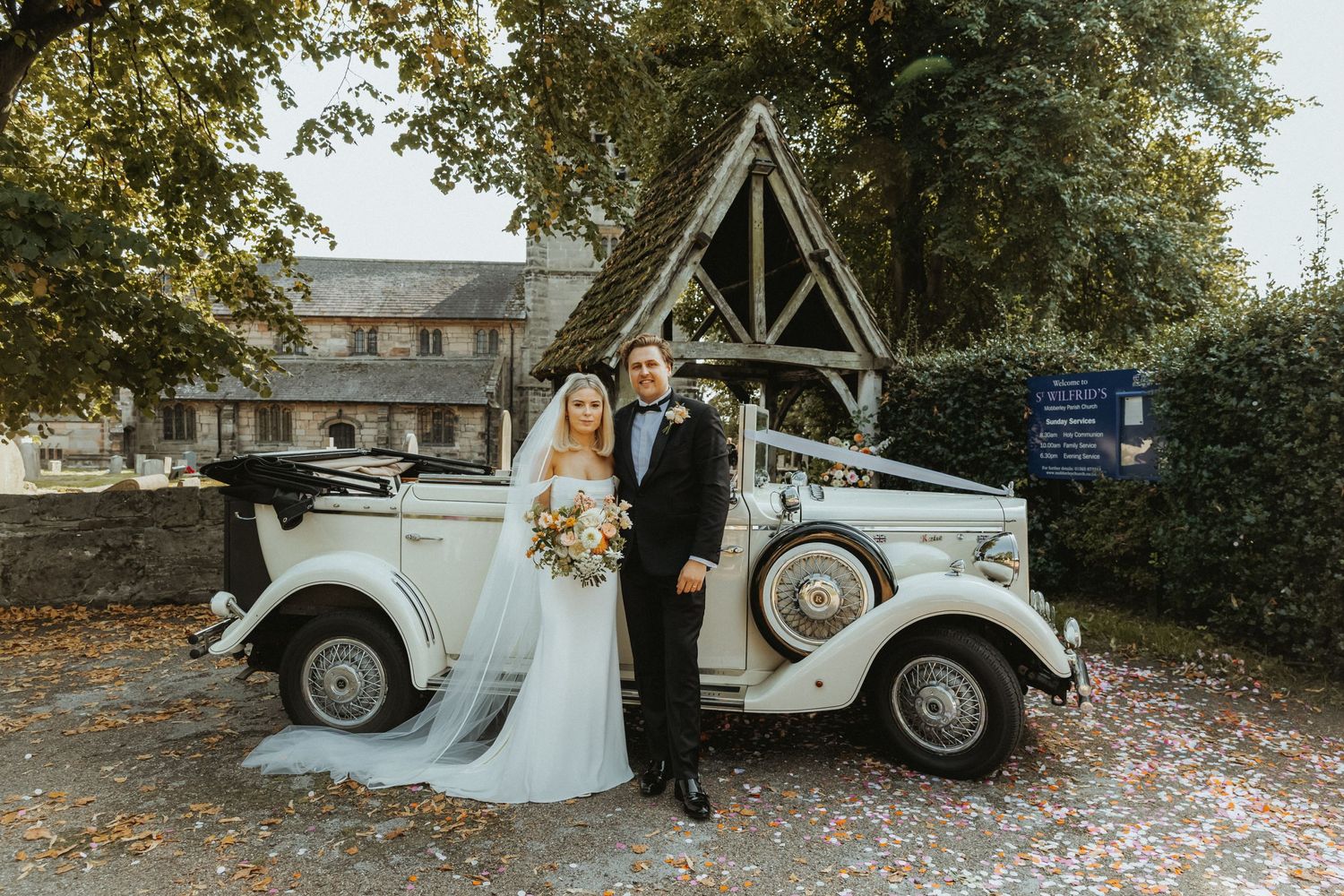 Bride and groom standing together next to vintage wedding car outside St Wilfrids church in Mobberley