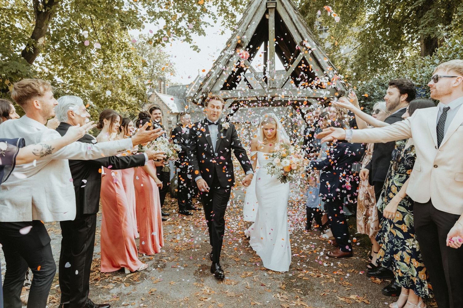Bride and groom walking through line of friends and family smiling and throwing confetti outside St Wilfrids church
