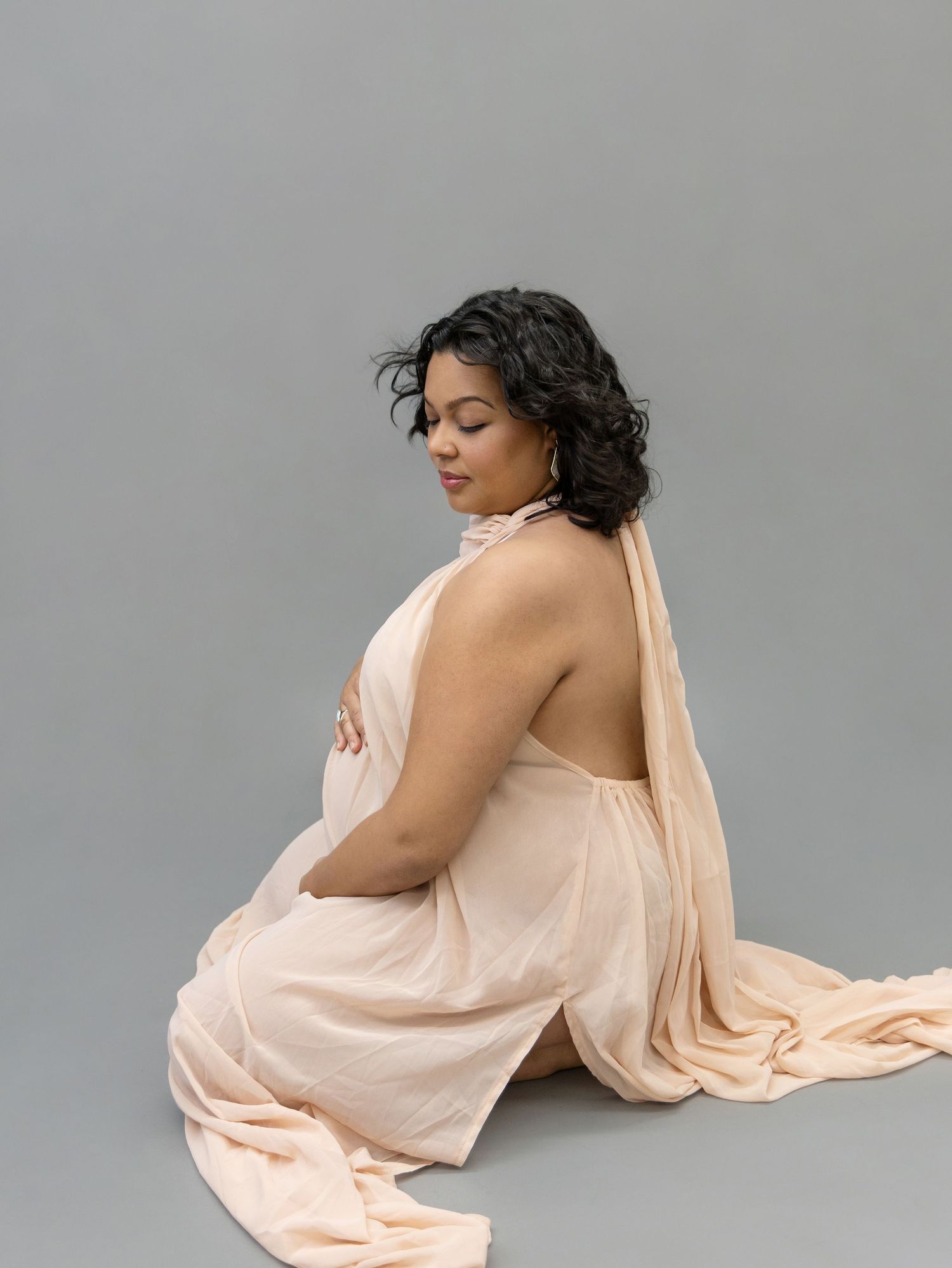Pregnant woman in elegant peach draped gown sitting gracefully on gray studio floor.