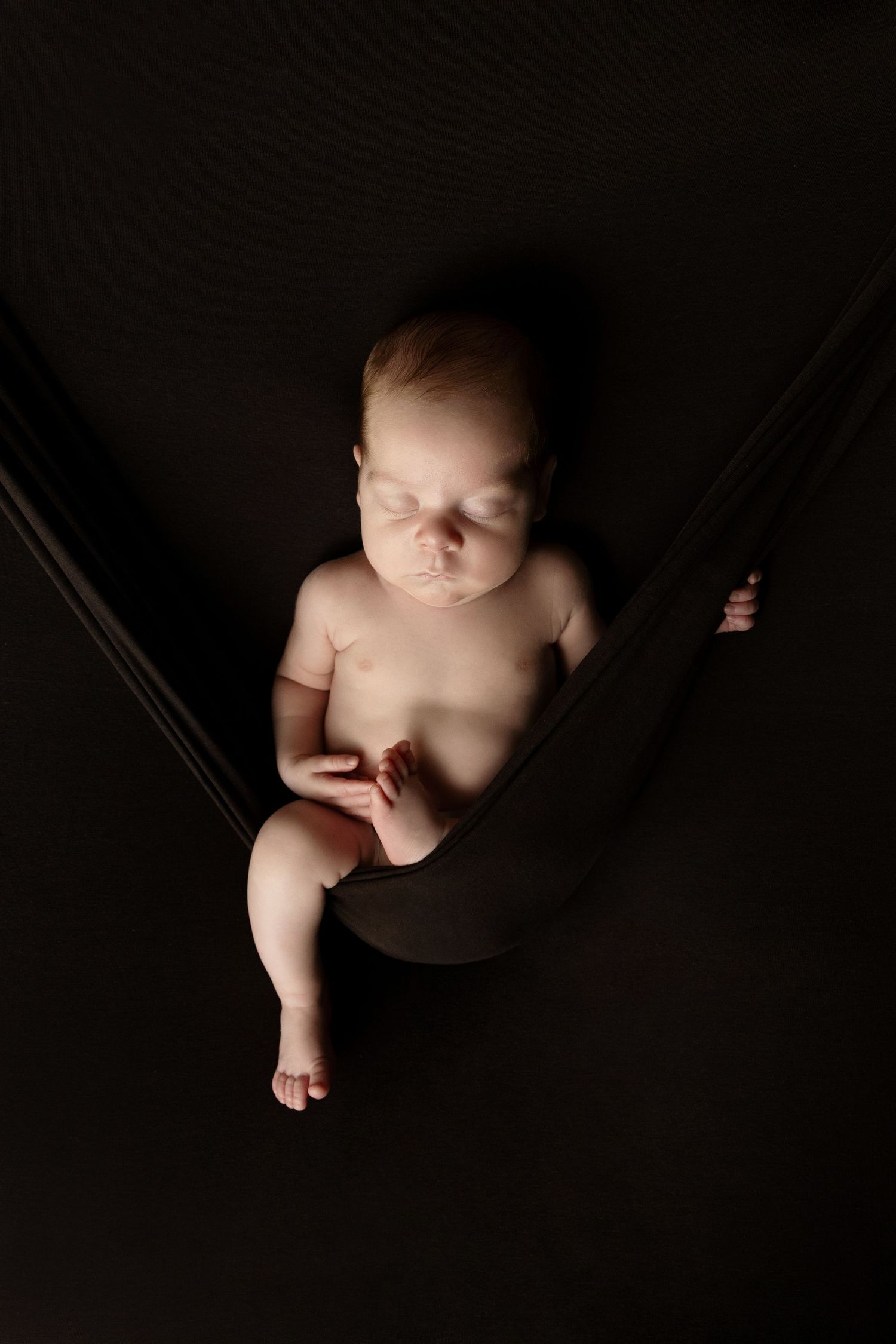 Newborn baby sleeping in a fabric hammock sling against a dramatic dark black background.