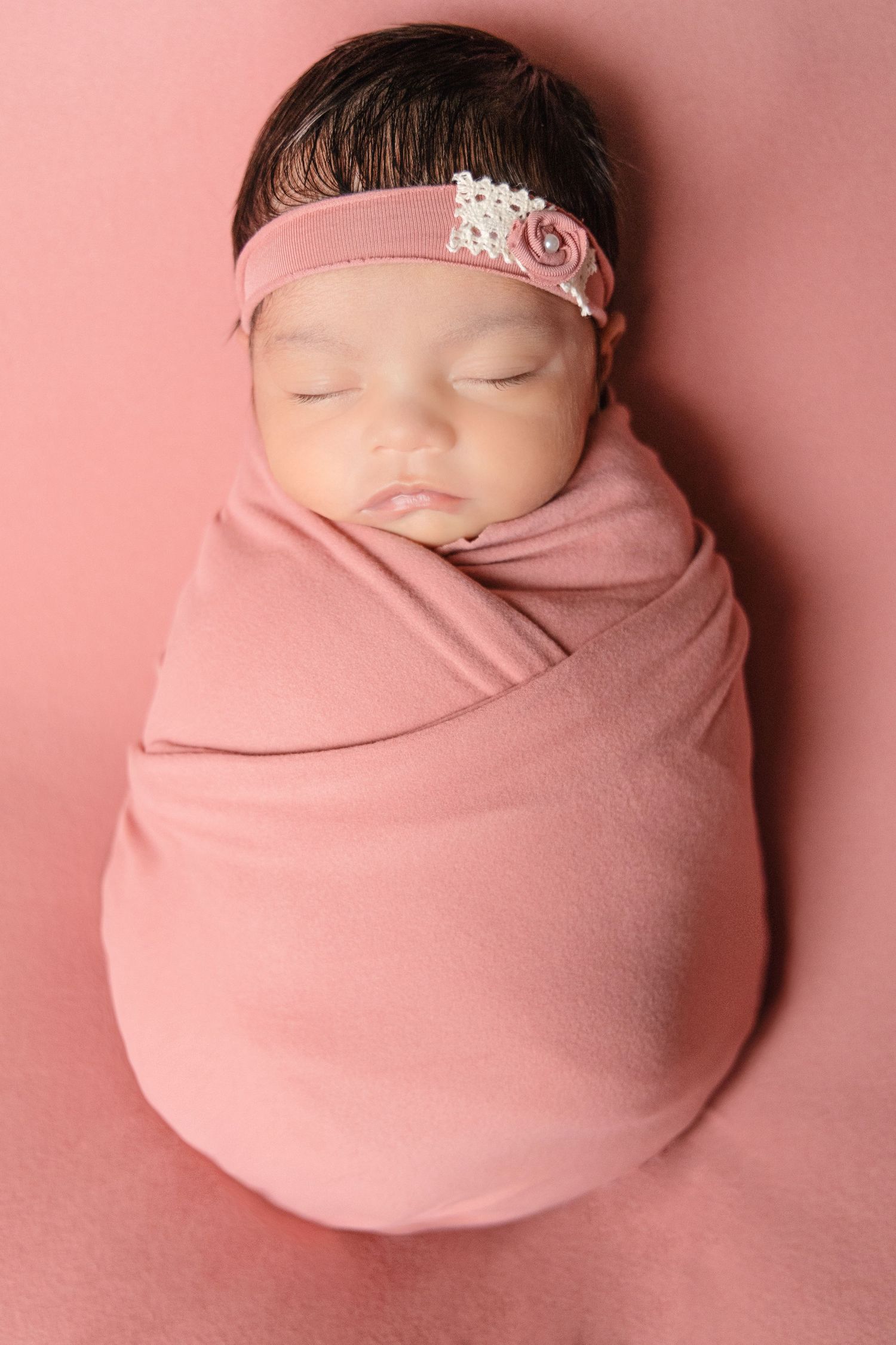 Newborn baby girl swaddled in pink wrap with floral headband, sleeping peacefully on pink background.