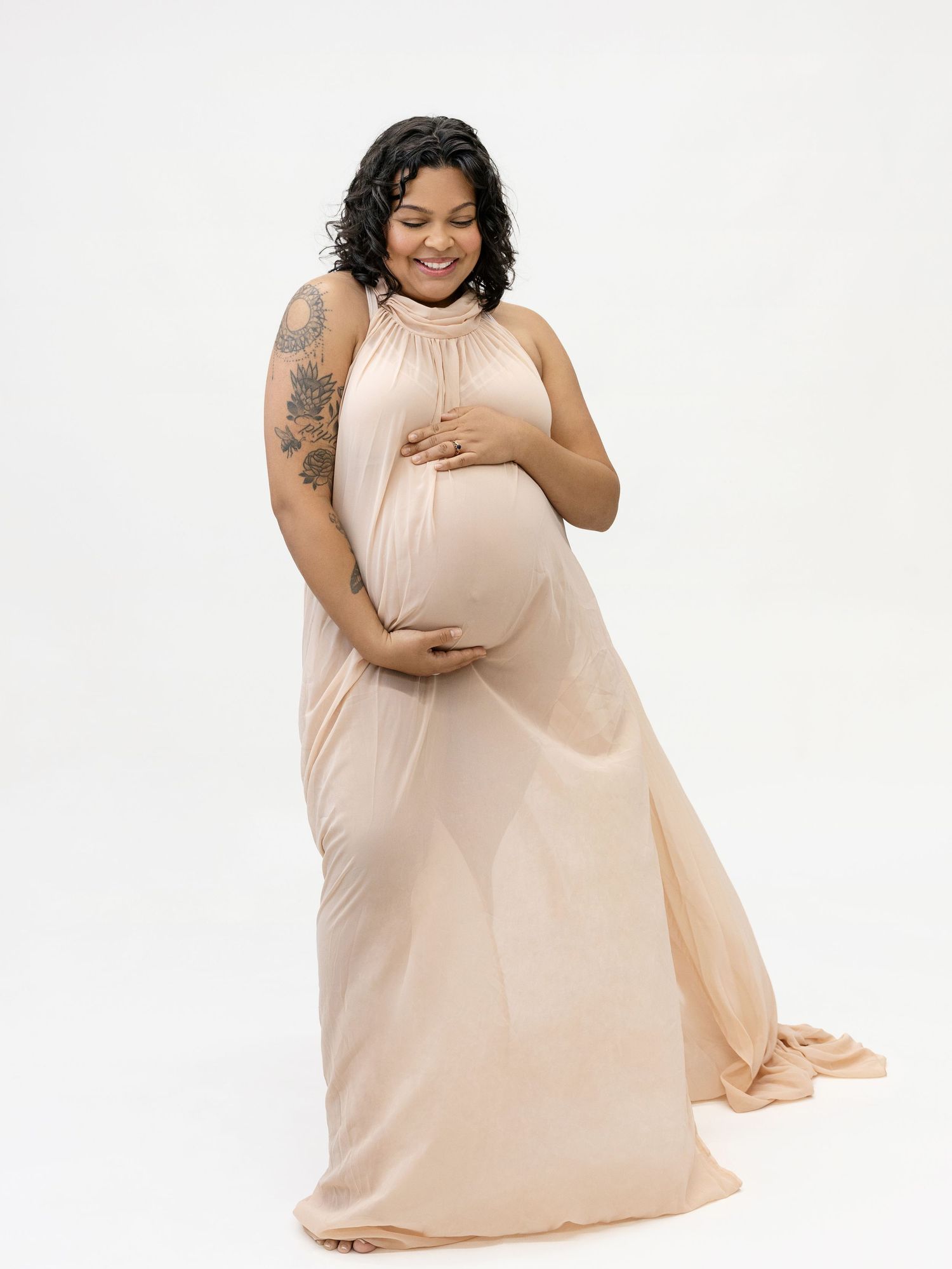 Pregnant woman in elegant peach halter maternity gown cradling baby bump, smiling downward in studio portrait.