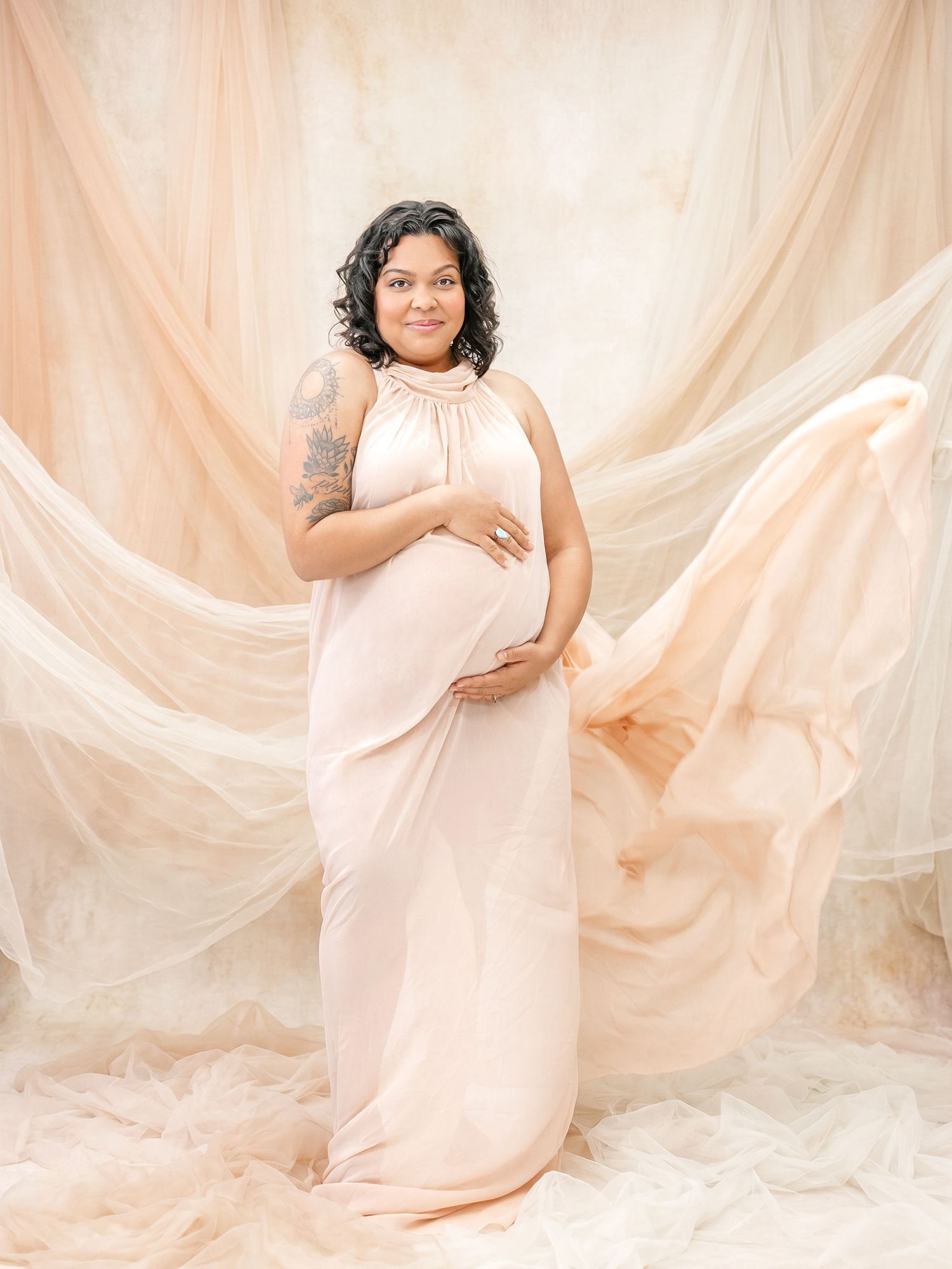 Pregnant woman in peach halter gown posing gracefully against flowing fabric backdrop in elegant studio maternity portrait.