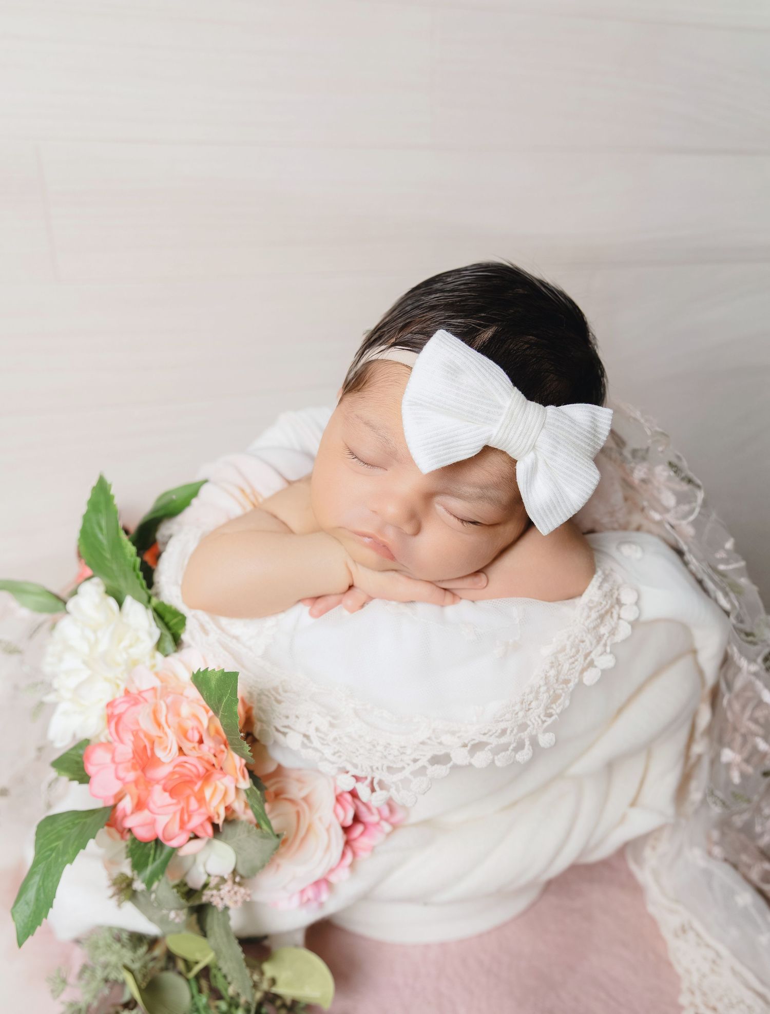Sleeping newborn baby girl in white lace wrap with bow beside flowers.