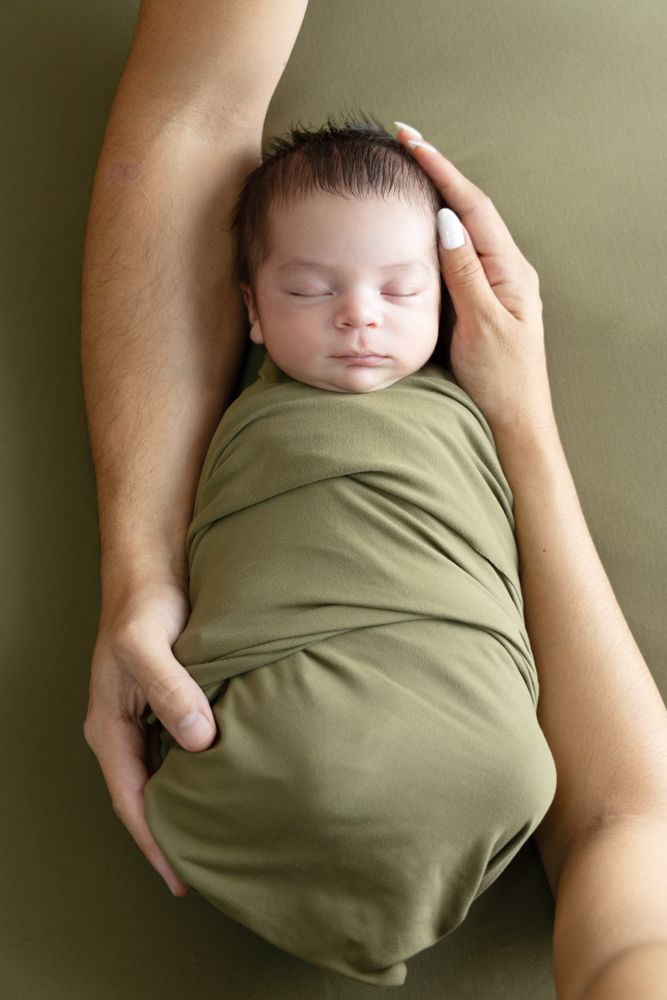 Newborn baby swaddled in olive green wrap sleeping peacefully while parent gently cradles and strokes head.