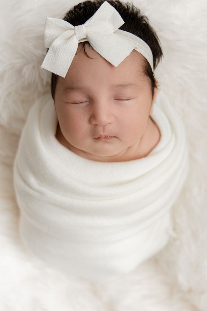 Sleeping newborn baby girl swaddled in white wrap with white bow headband on soft white fur blanket.