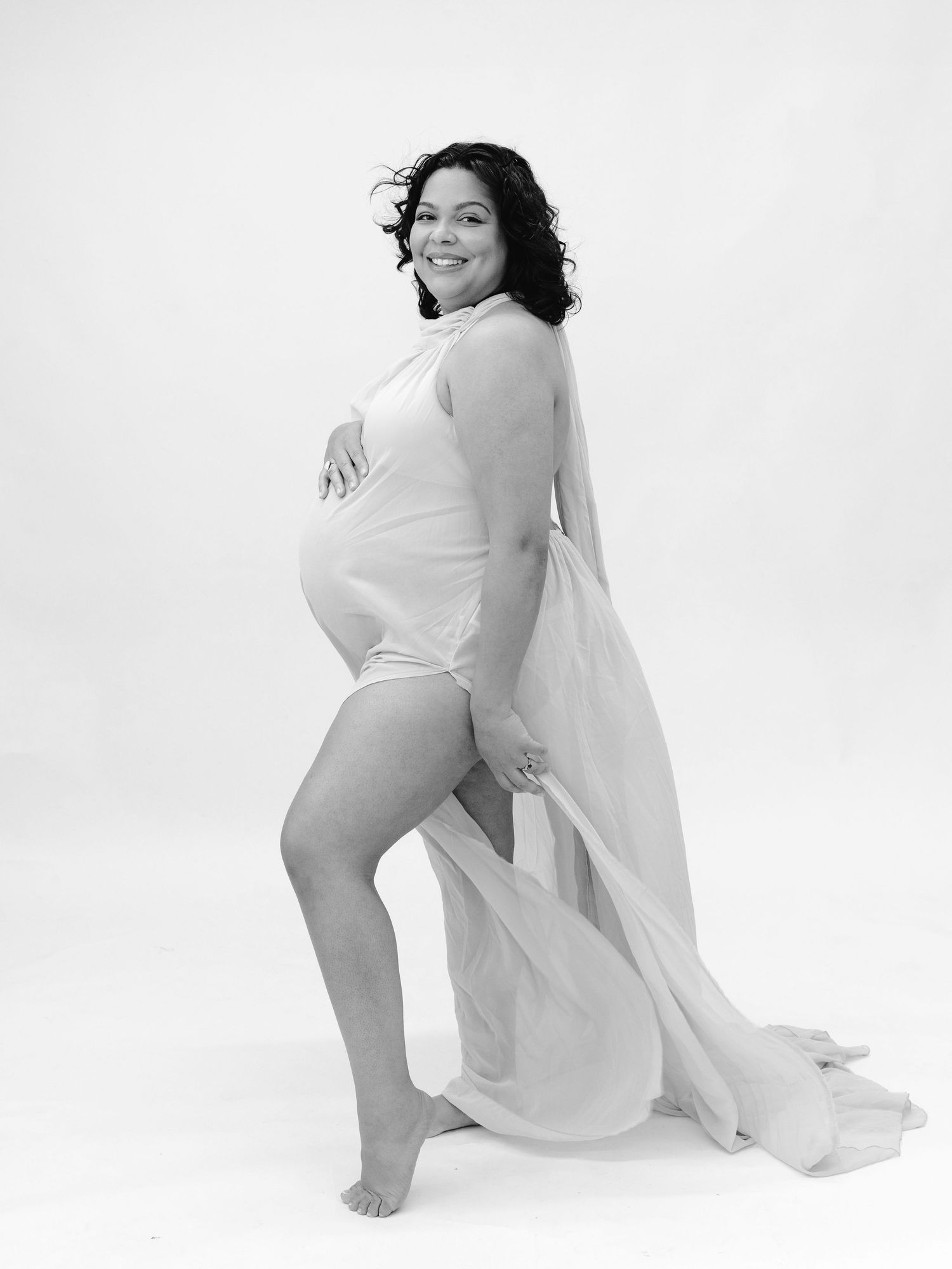 Black and white maternity portrait of smiling pregnant woman in sheer white fabric wrap, posing sideways in studio.