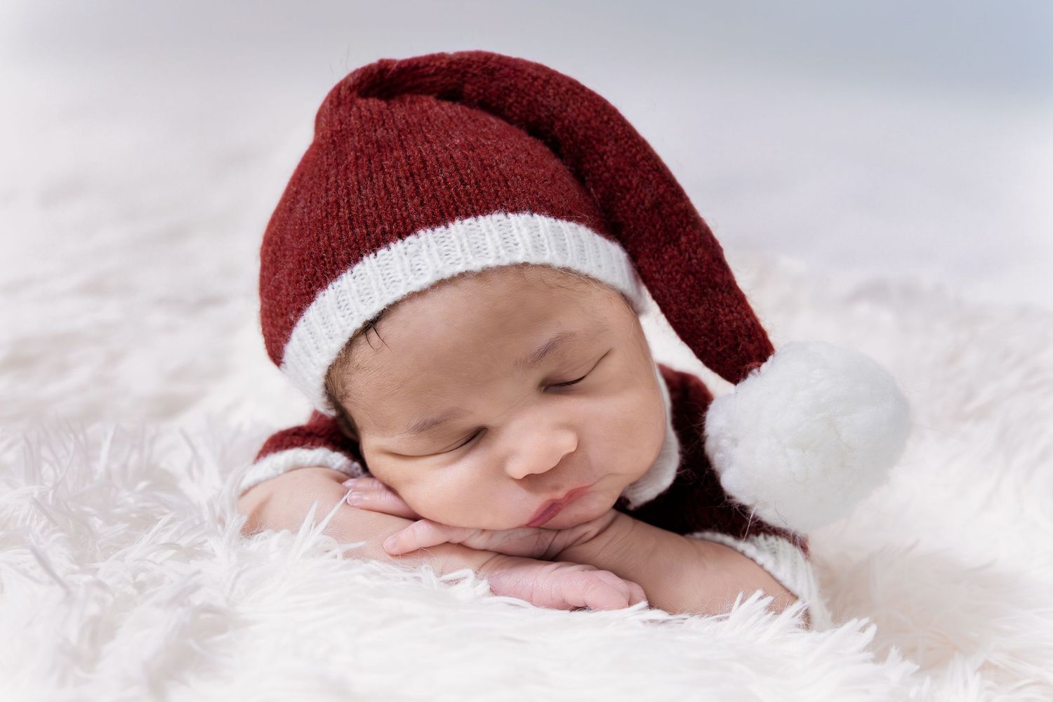 Adorable newborn baby sleeping with chin on hands wearing a red and white Santa knit hat.