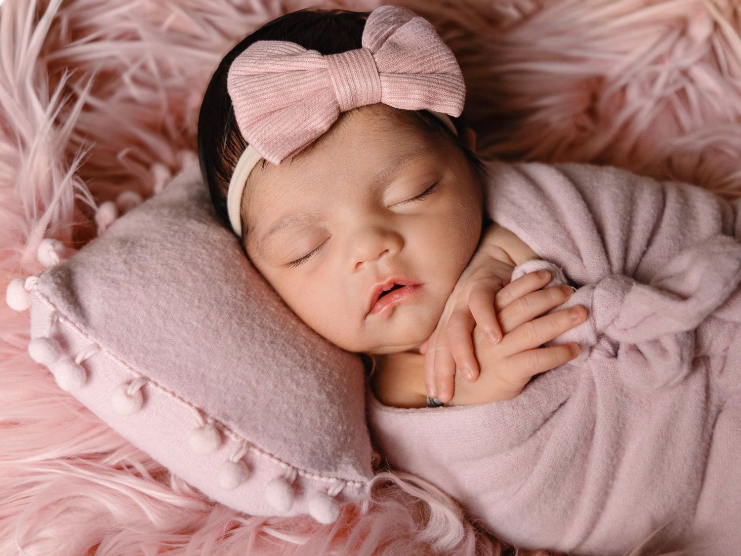 Newborn baby girl sleeping on pink pillow with bow headband wrapped in soft pink blanket.