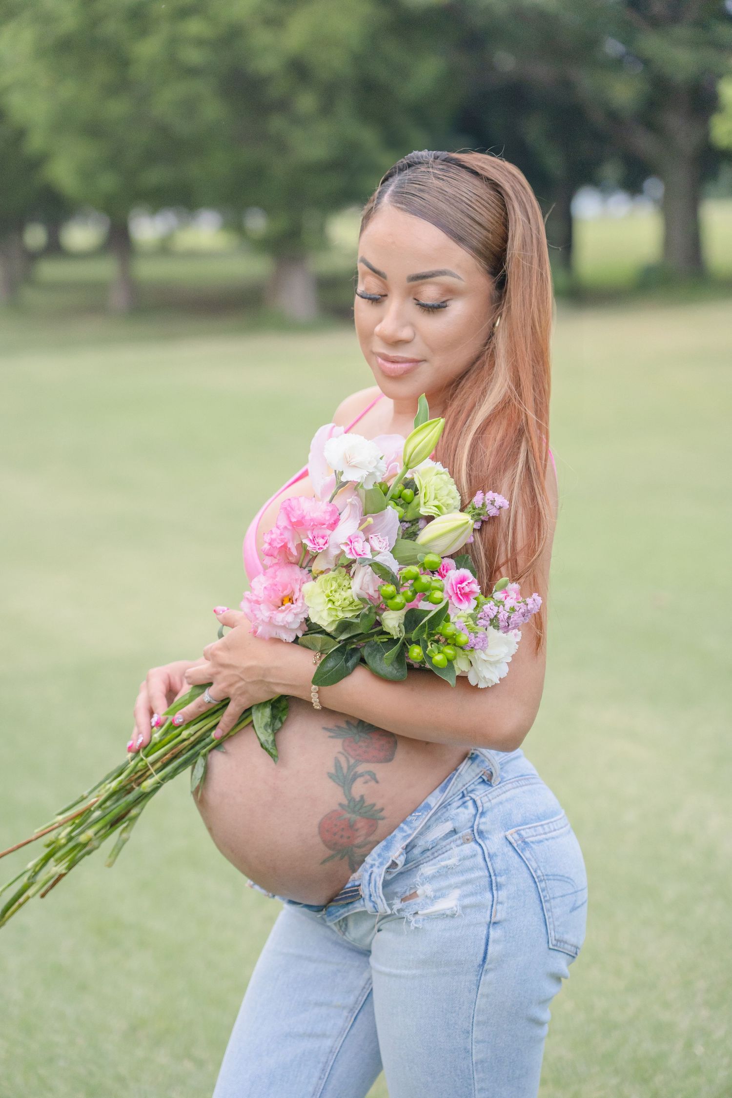Pregnant woman with long hair holding pink and white flower bouquet, showing tattooed baby bump in outdoor park setting.