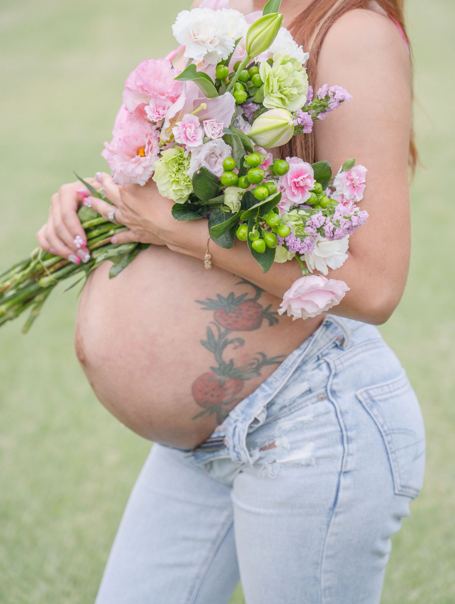 Close-up of pregnant belly with floral tattoo, woman holding bouquet of pink and green flowers outdoors.