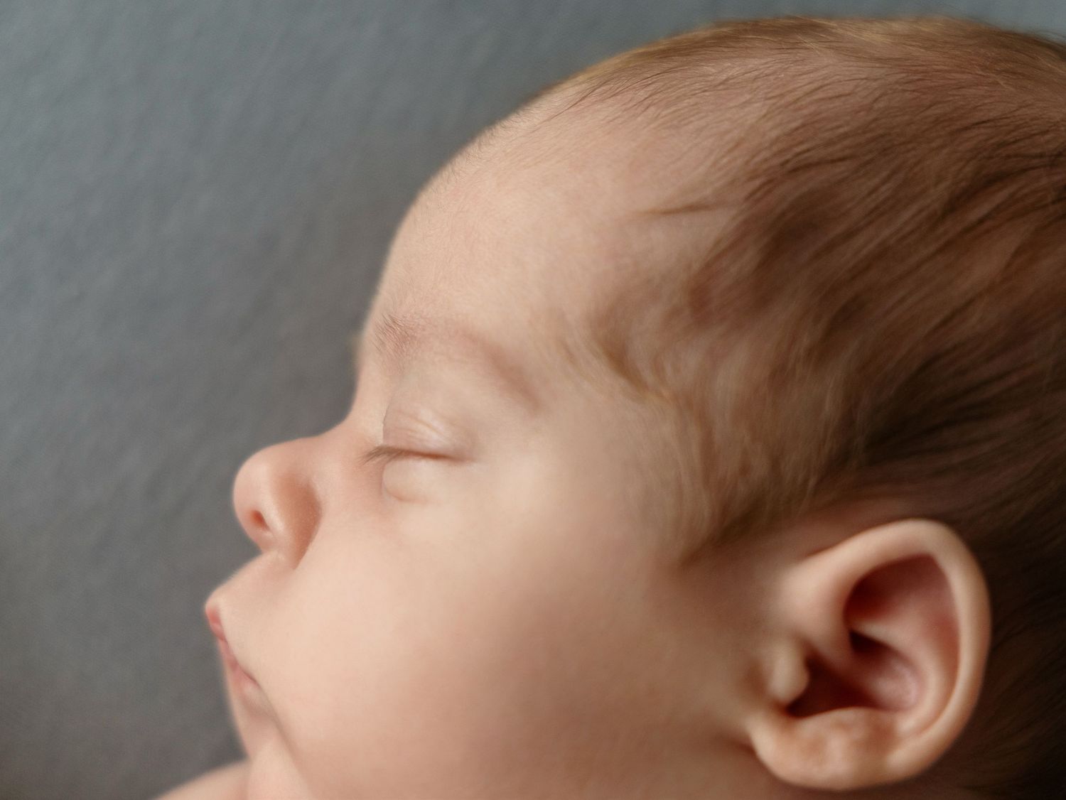 Close-up side profile of a sleeping newborn baby with soft features on gray background.