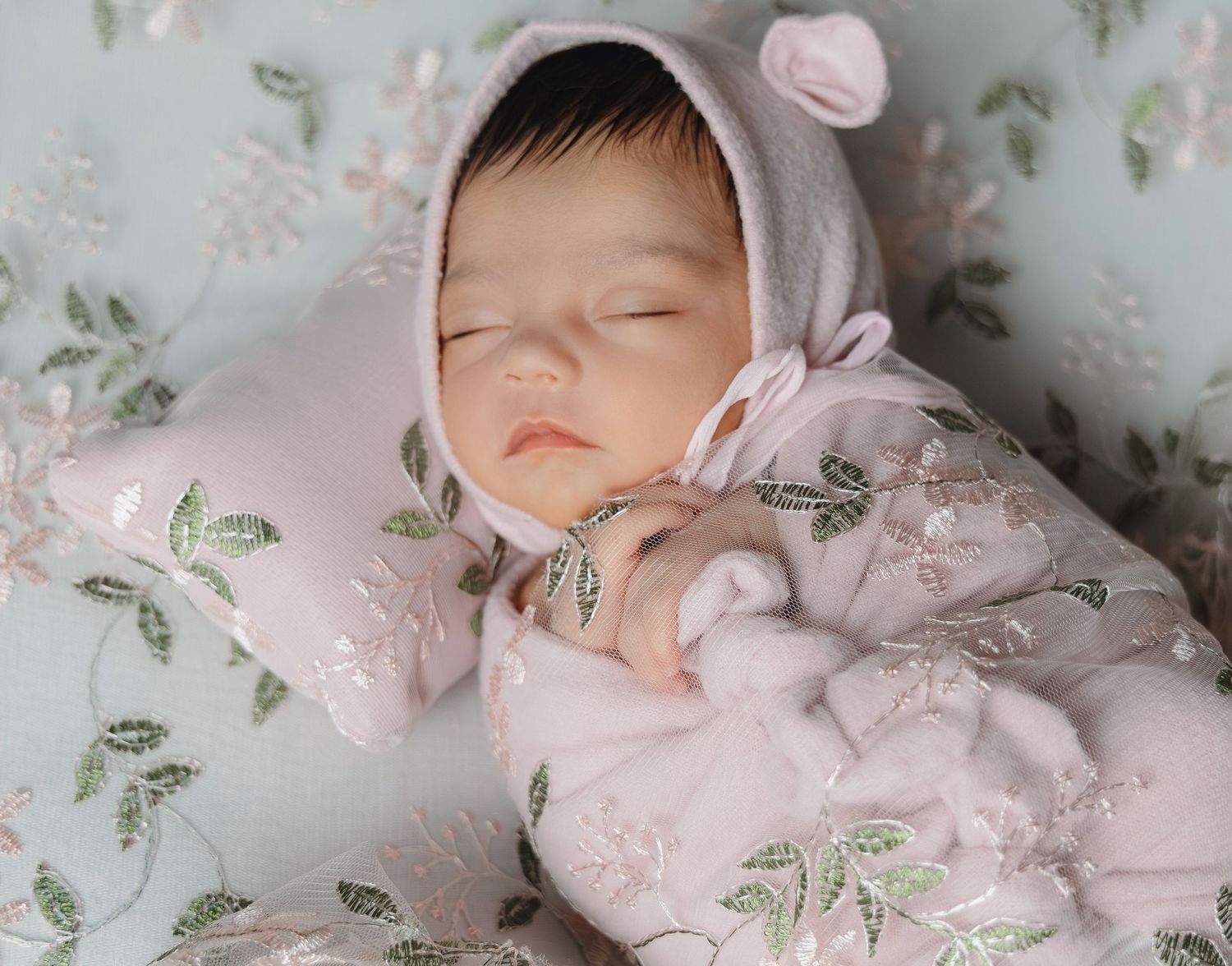 Sleeping newborn baby in pink floral outfit and bear bonnet hat resting on matching embroidered pillow.