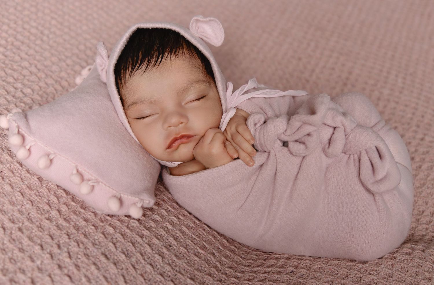 Sleeping newborn baby wrapped in a pink bunny outfit with ear bonnet on a knit blanket.