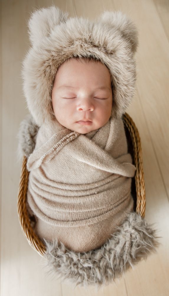 Sleeping newborn baby swaddled in beige wrap wearing fuzzy bear ear bonnet, nestled in wicker basket.
